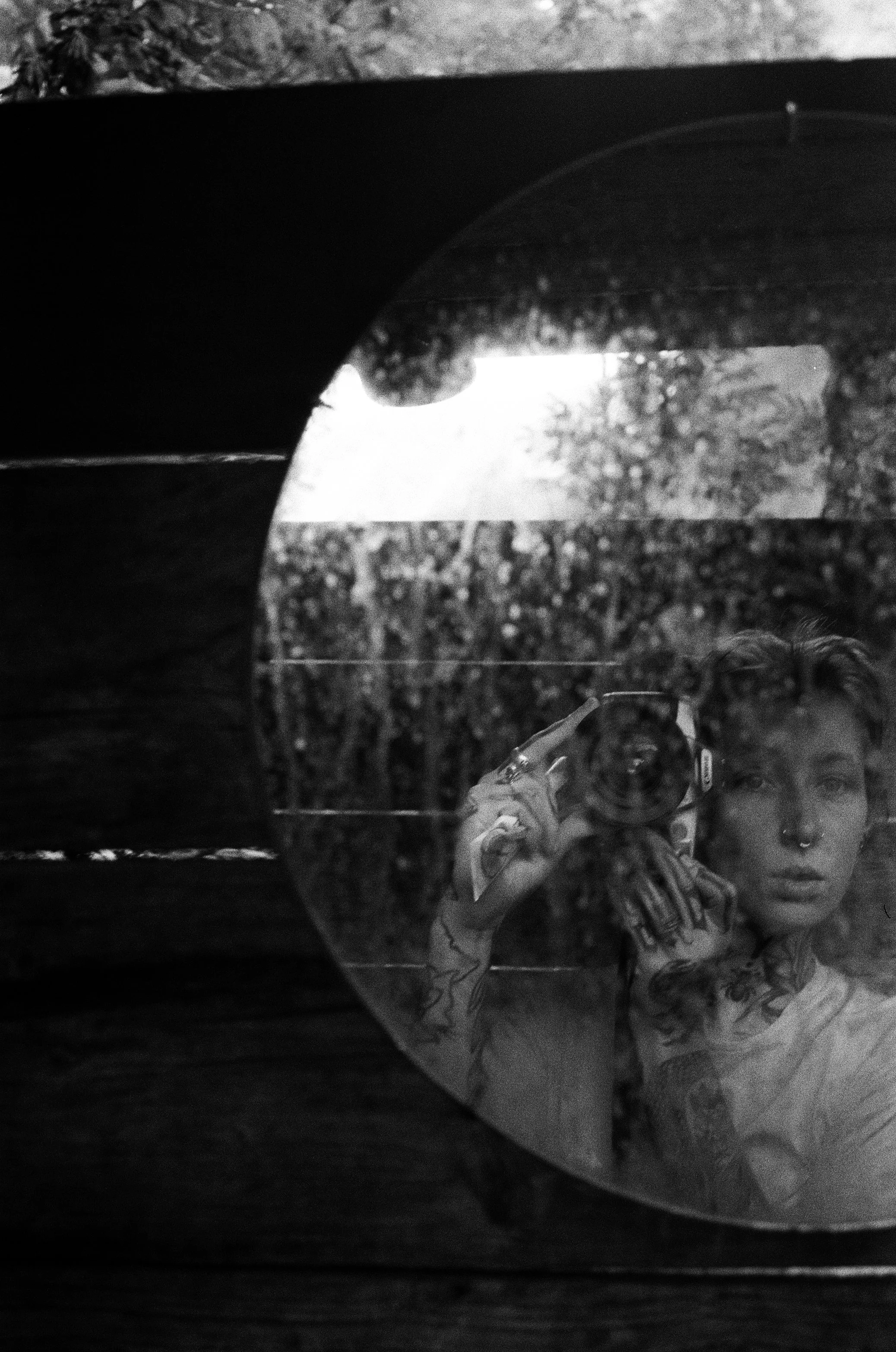 Reflet d'une personne avec des tatouages, tenant un appareil photo, pris dans une surface en verre ou en miroir, avec un fond de végétation ou d'extérieur, en noir et blanc.