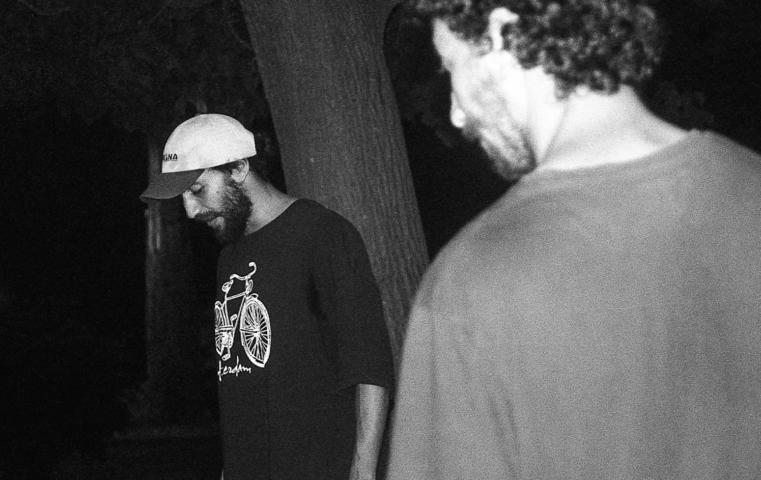 Deux hommes au coucher du soleil ou dans la nuit, un portant un t-shirt noir avec un dessin de vélo et un chapeau, l'autre portant une chemise claire et des lunettes, près d'un arbre.