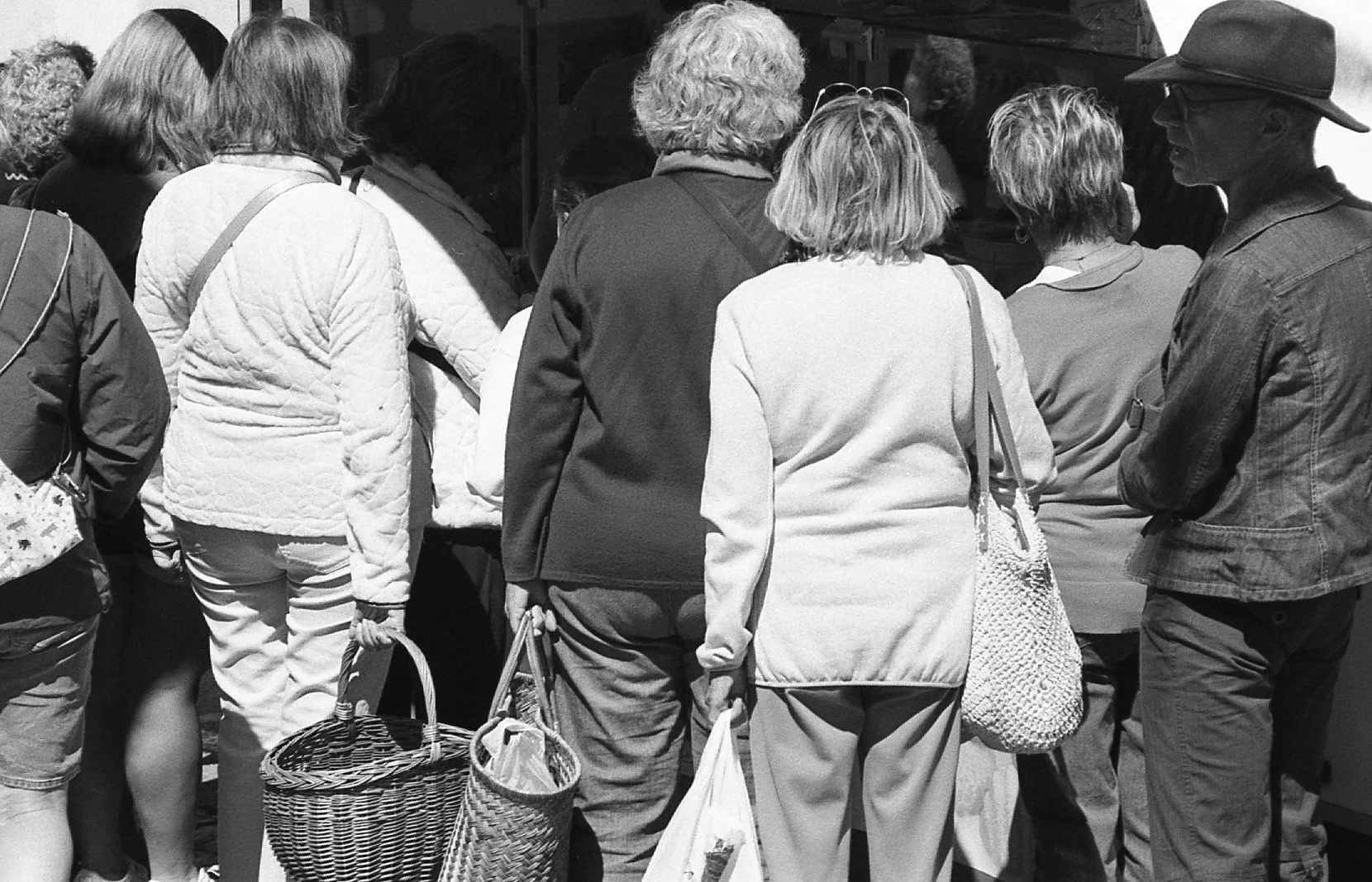 Groupe de personnes faisant la queue, photographiées en noir et blanc, vues de dos, portant des vêtements décontractés et des sacs