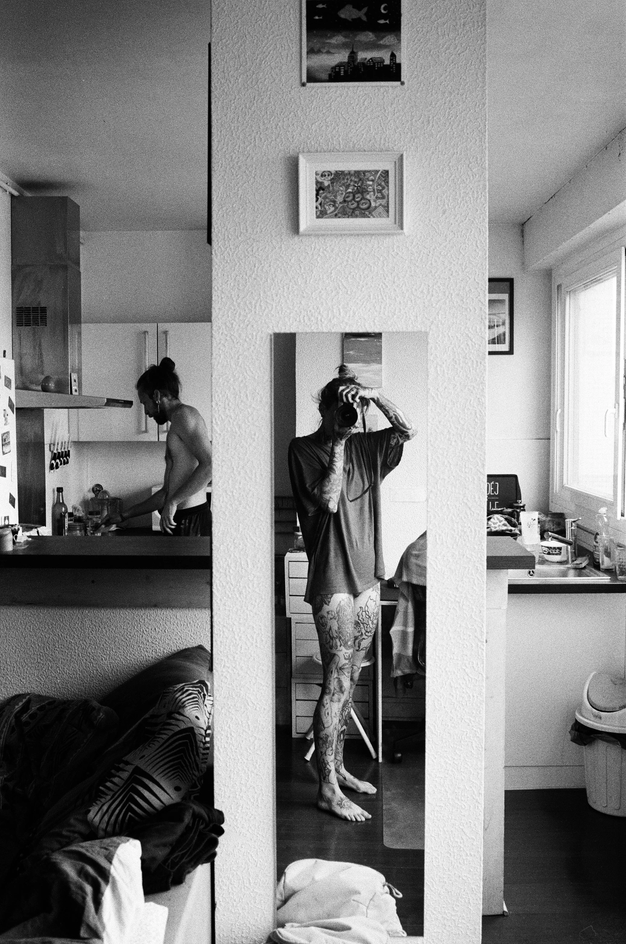 Photo en noir et blanc d'une femme avec des tatouages, se tenant devant un miroir, tenant un appareil photo, dans un salon, avec divers objets et meubles visibles.