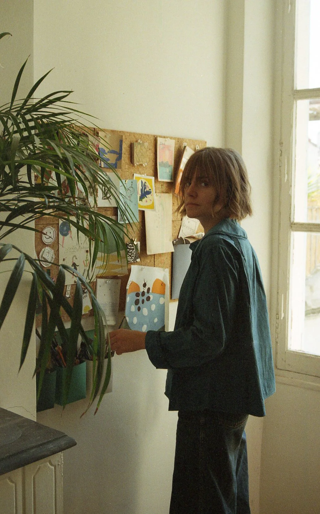 Une jeune femme à cheveux bruns courts, portant une veste en jean foncé, se tient près d'un tableau d'affichage avec des dessins et des cartes, dans une pièce lumineuse avec une fenêtre et un mur blanc.