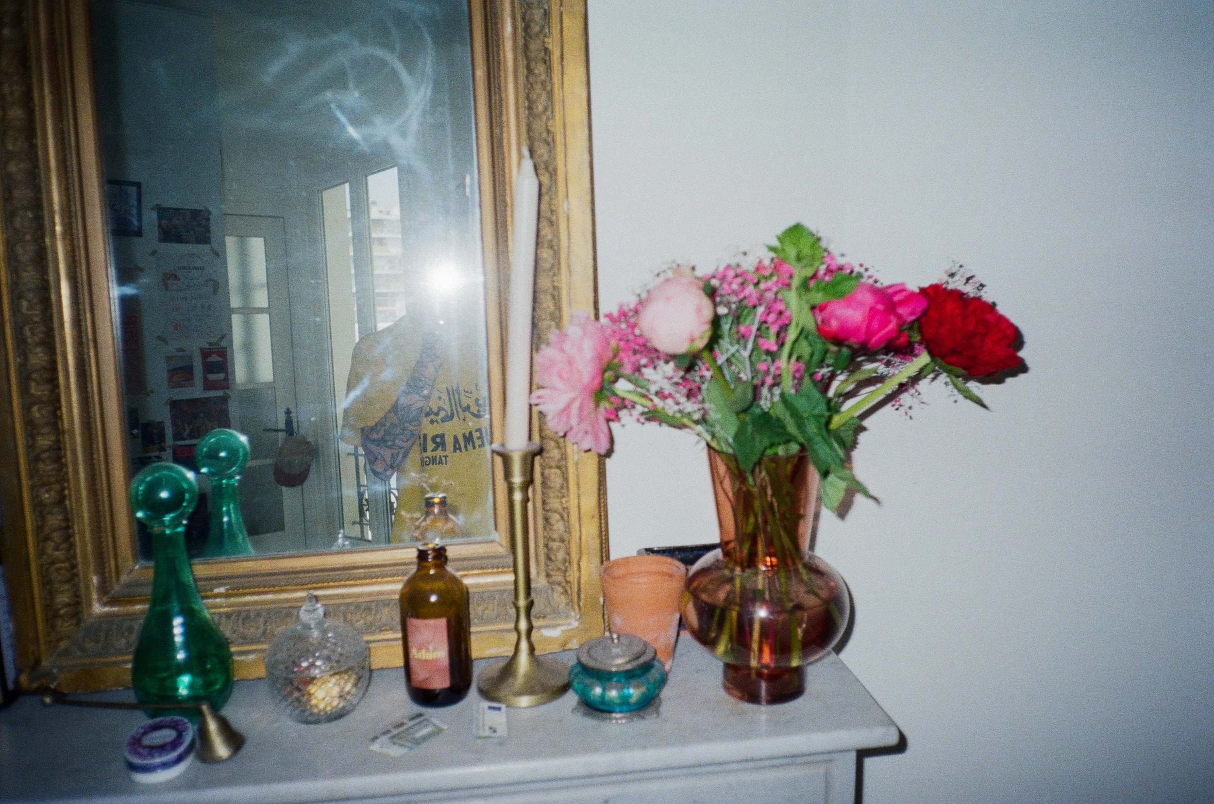 Un miroir ancien orné de détails dorés, une table avec plusieurs bouteilles en verre, un vase avec un bouquet de fleurs roses et rouges, ainsi que divers objets décoratifs en verre et en métal.