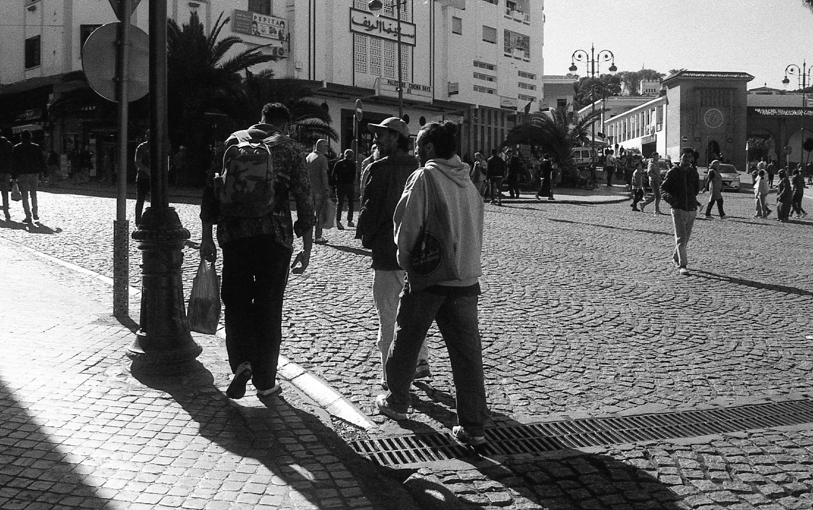 Rue animée avec plusieurs personnes marchant, bâtiments en arrière-plan, lampadaires, et un sol pavé, scène en noir et blanc