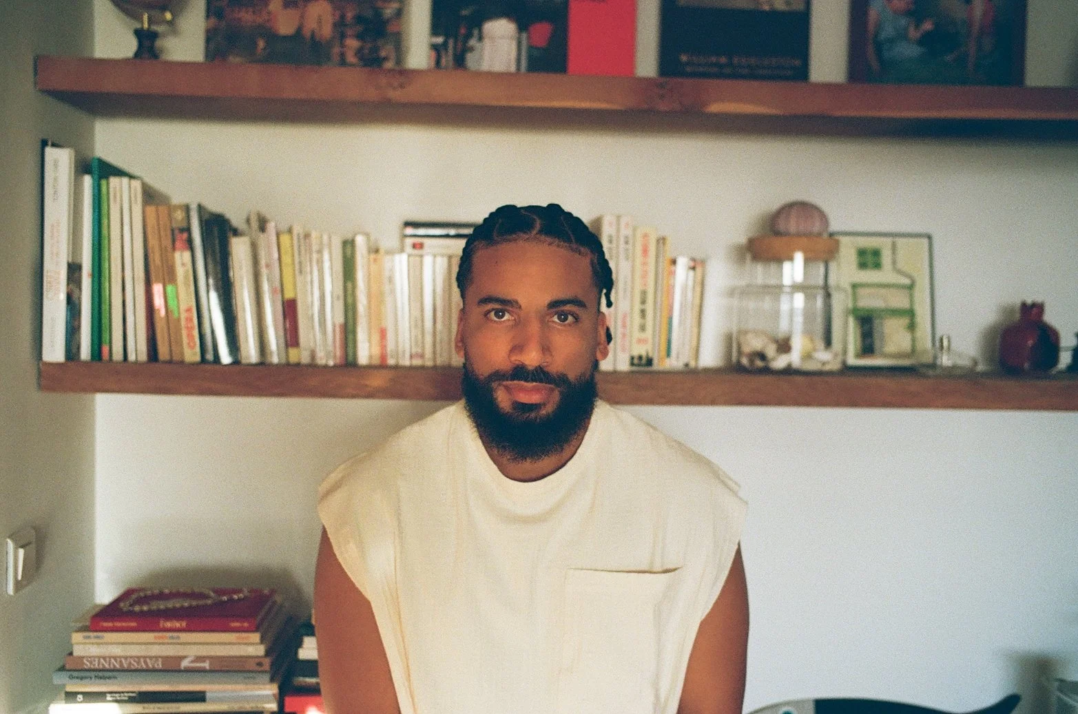 Un homme avec une barbe et des cheveux tressés, portant une tenue beige, assis devant une bibliothèque remplie de livres.