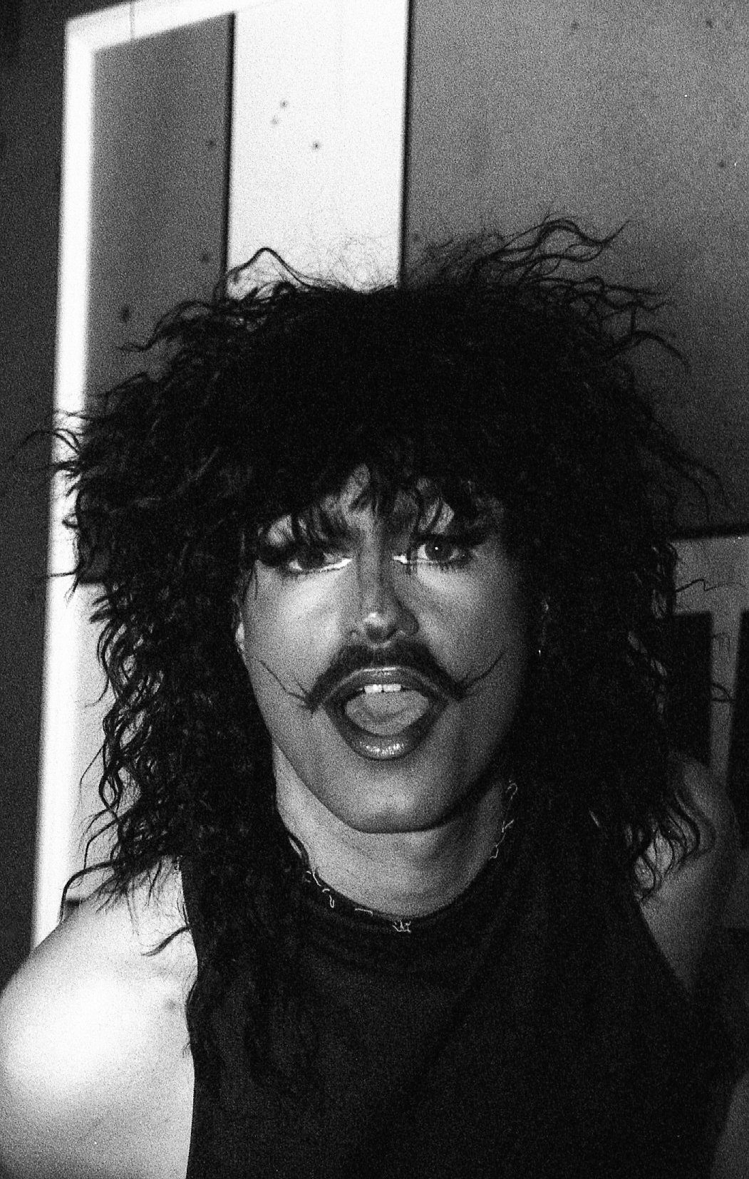 Une personne avec des cheveux bouclés et un maquillage dramatique pose devant un mur. La photo est en noir et blanc.