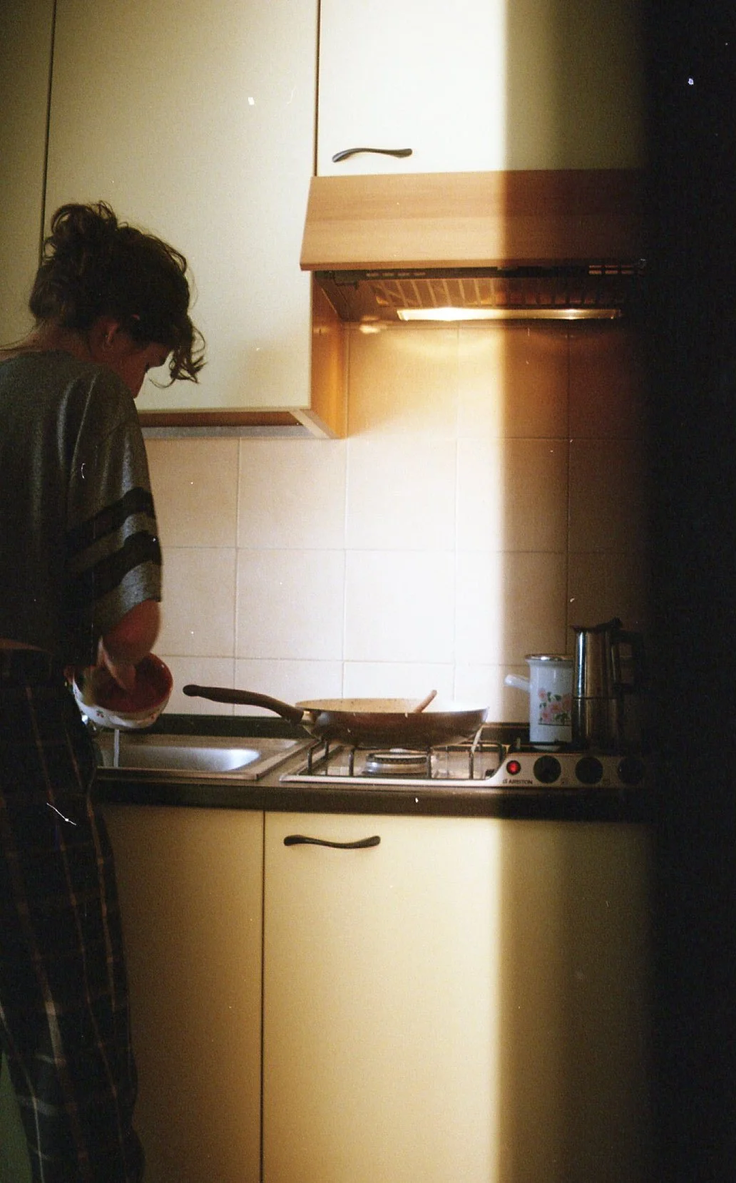 Une femme qui cuisine dans une petite cuisine, avec un évier, une cuisinière à gaz, et des placards en bois.