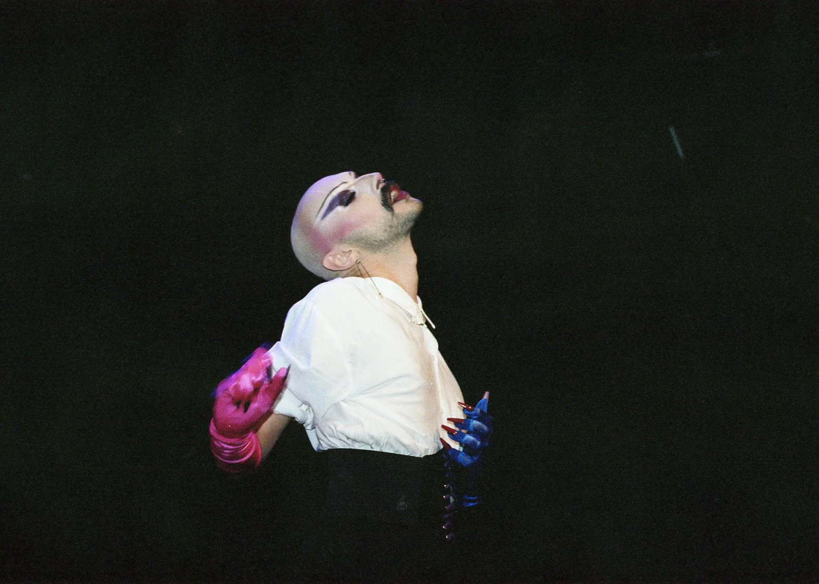 Une personne avec maquillage extravagant, portant une chemise blanche et des gants colorés, pose avec la tête levée contre un fond noir.
