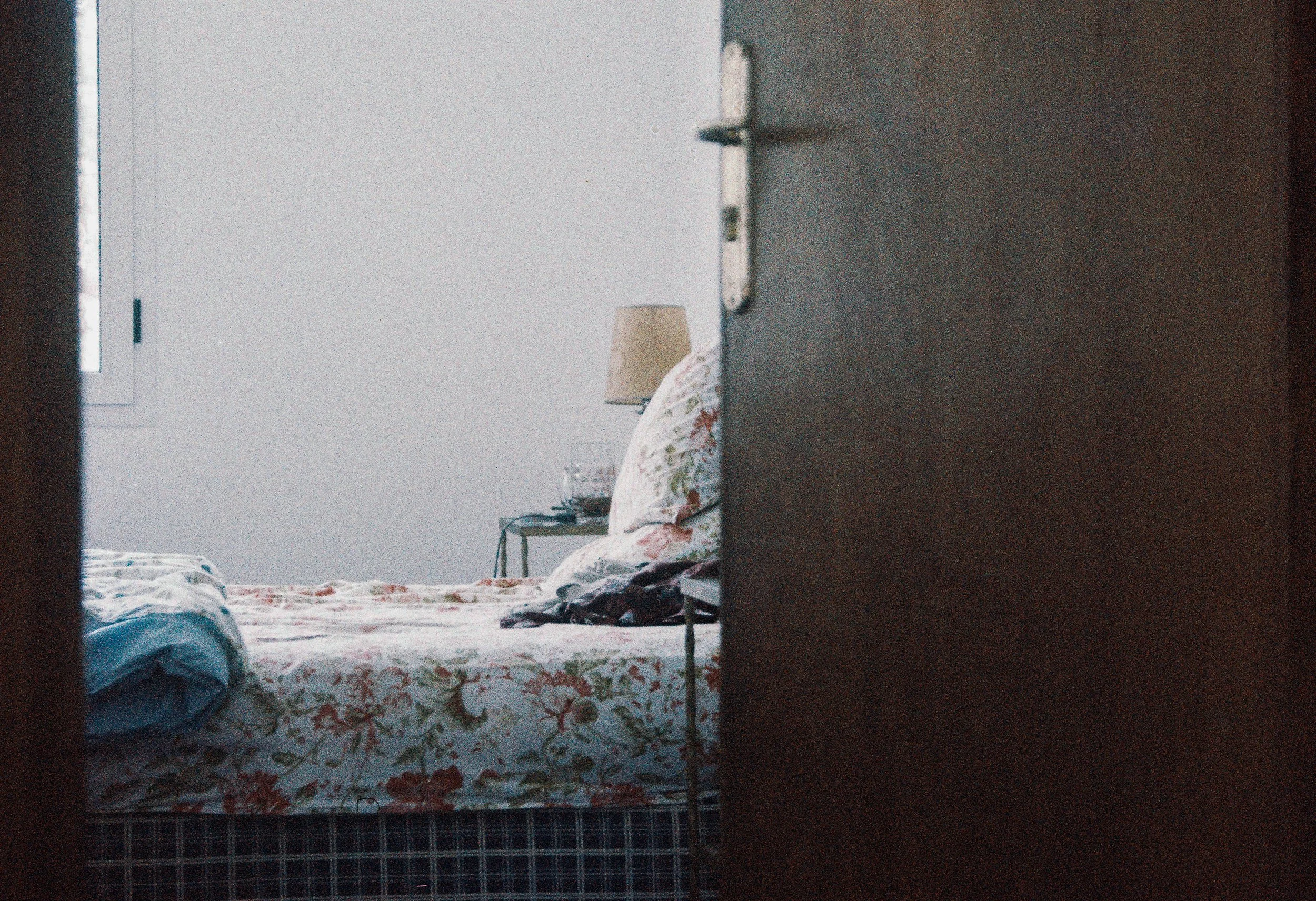 Vue partielle d'un lit avec literie à motifs floraux, une table de nuit avec une lampe et un verre d'eau, vue à travers une porte entrouverte dans une pièce lumineuse