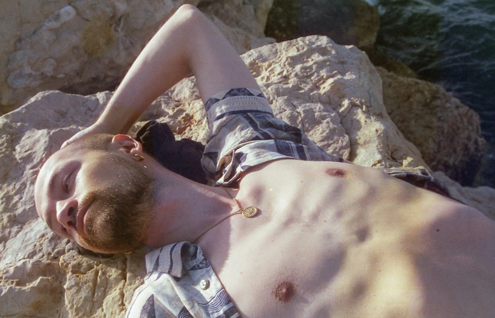 Un homme bronzant sur des rochers près de l'eau, portant une chemise à rayures légère ouverte et une chaîne avec un pendentif.