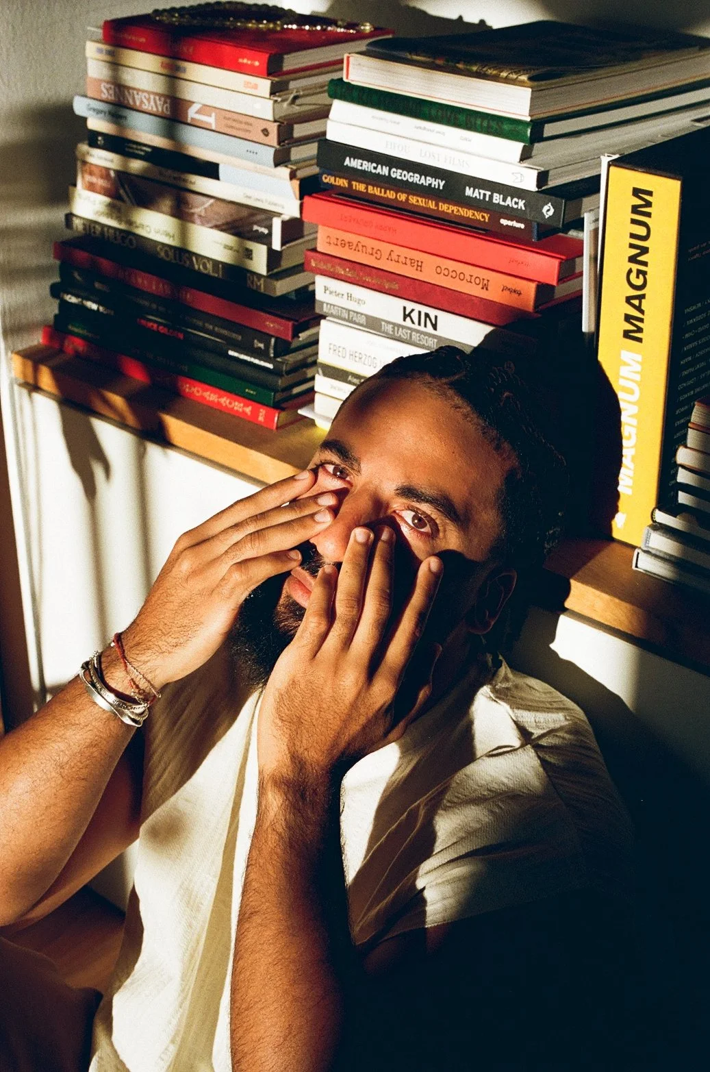 Un homme avec une barbe, portant un bracelet, est assis près d'une étagère remplie de livres. Il se touche le visage avec ses mains, regardant vers l'objectif.