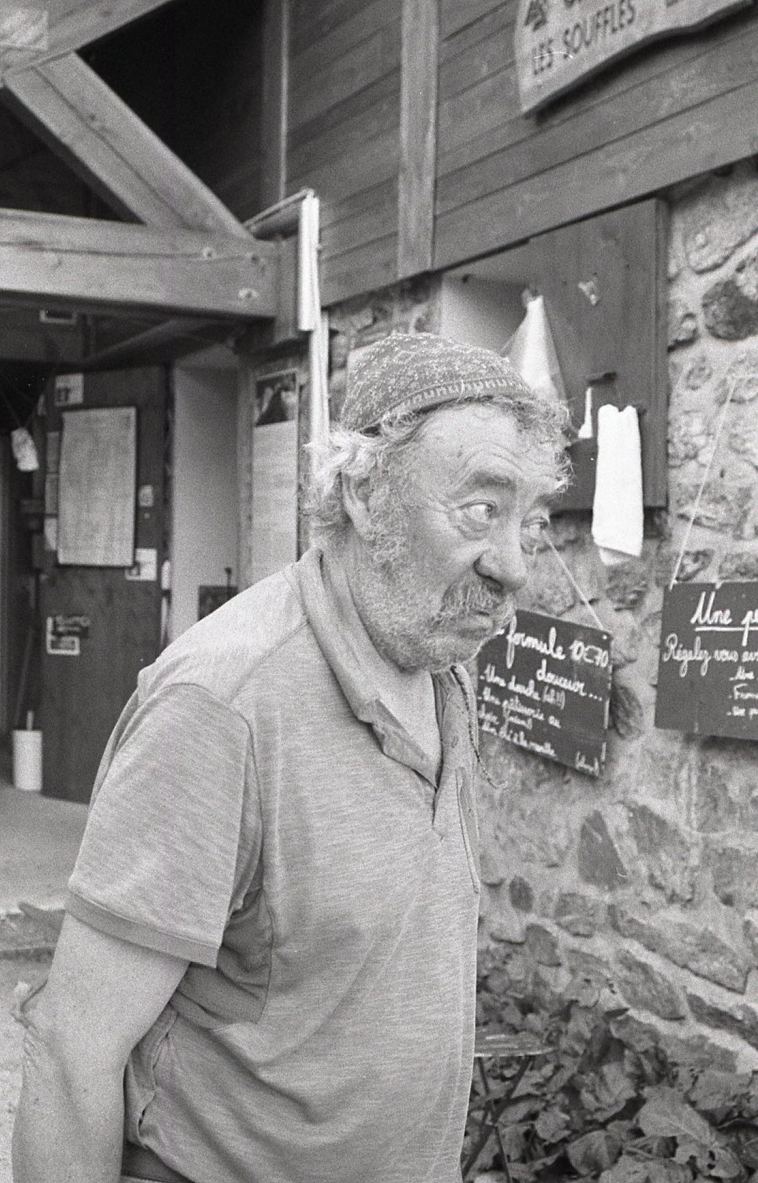 Un homme âgé avec une barbe et des cheveux gris, portant un chapeau, debout devant un mur en pierre avec des tableaux noirs et des papiers accrochés.