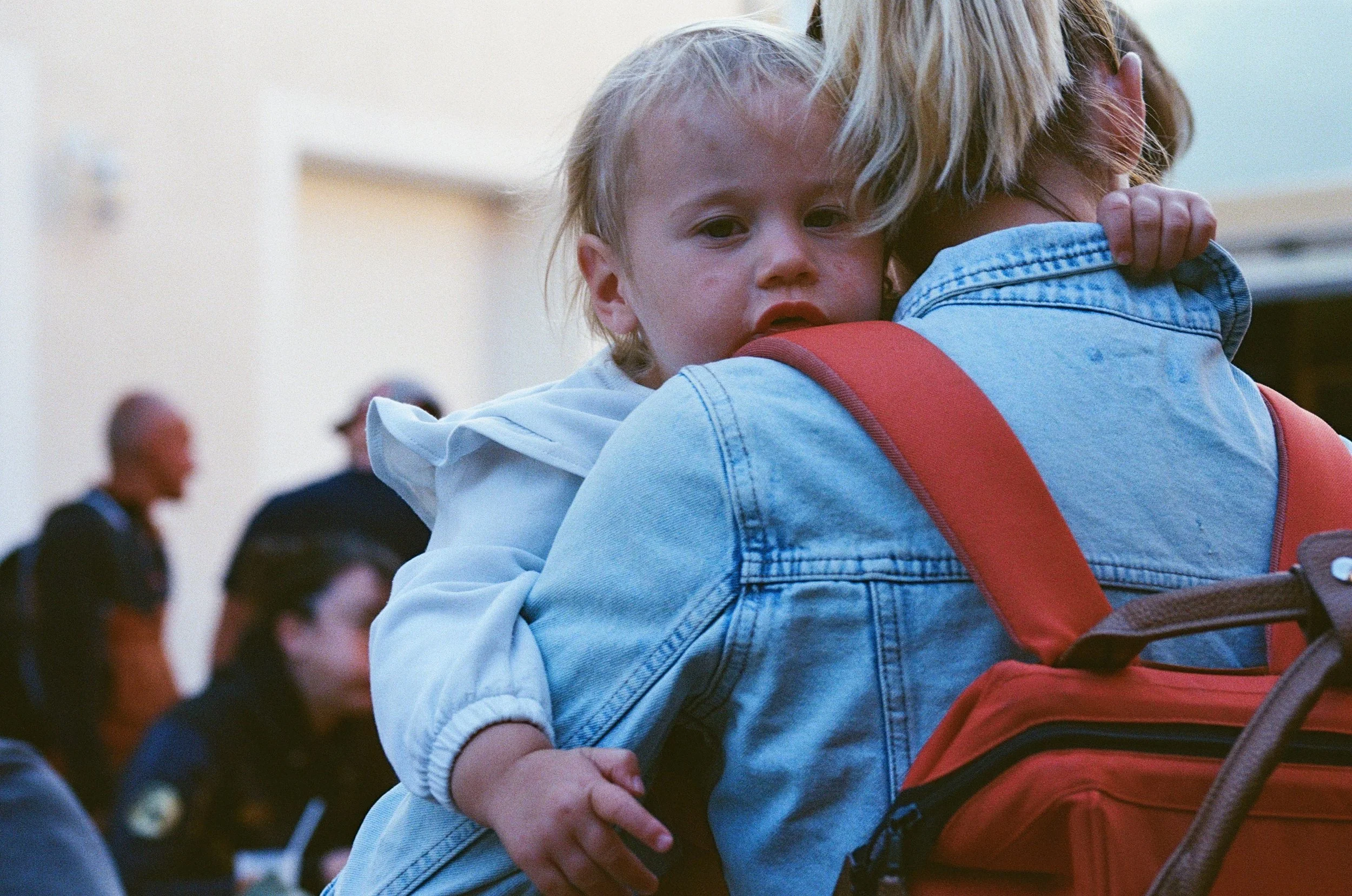 Un enfant fatigué ou triste est porté par une adulte, dans un lieu public avec d'autres personnes.