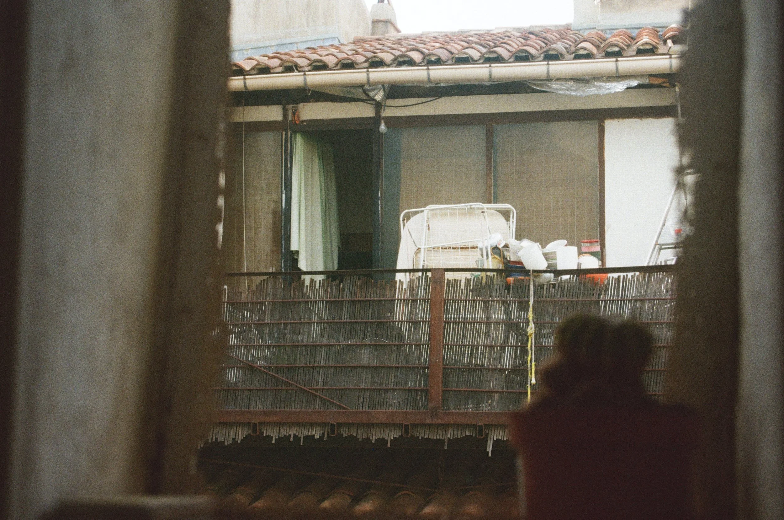 Vue d'un balcon d'un appartement avec des objets ménagers, encadrée par deux murs en pierre, présentant une toiture en tuiles.