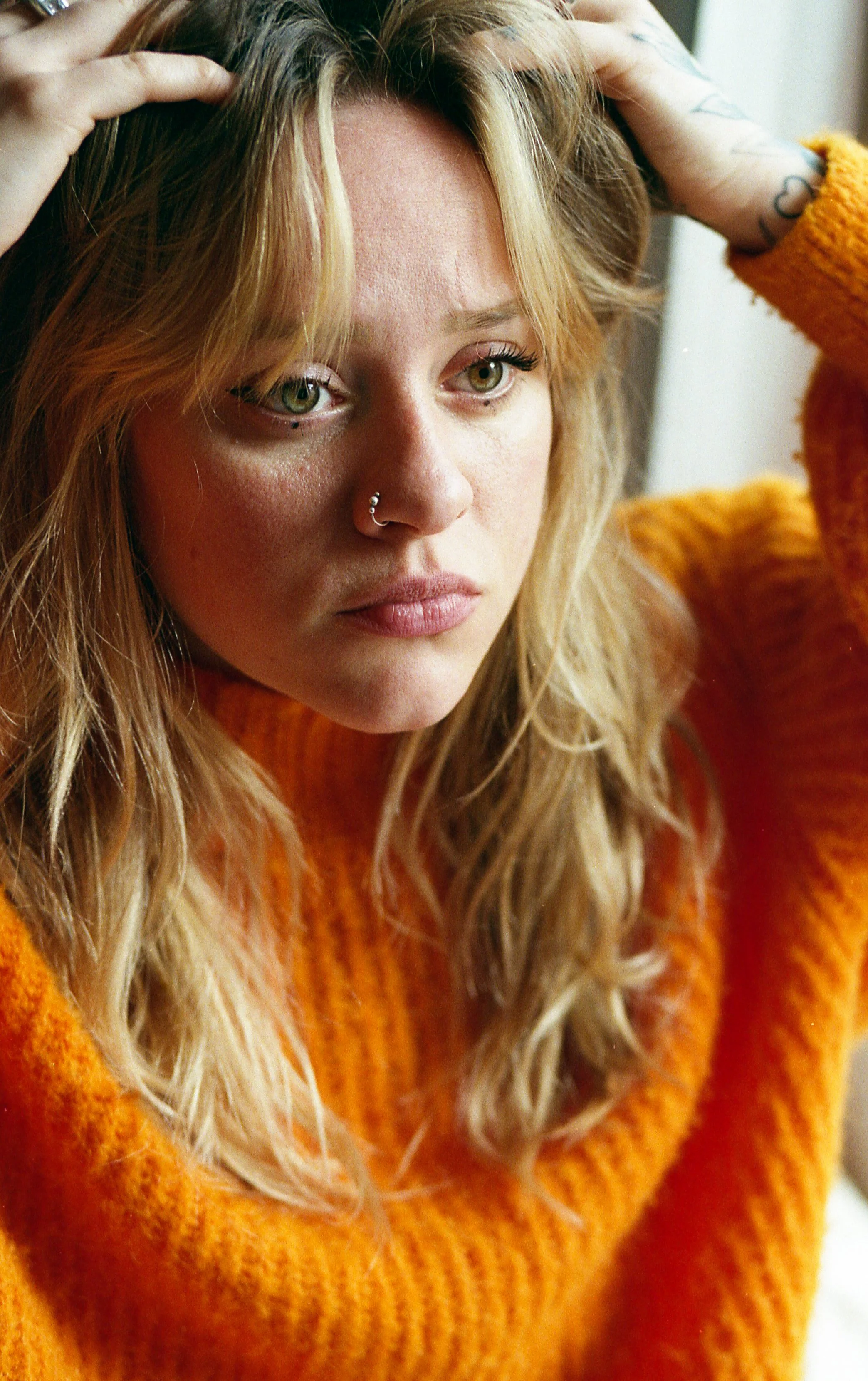 Une jeune femme aux cheveux blonds, portant un pull orange, pose en regardant pensivement.