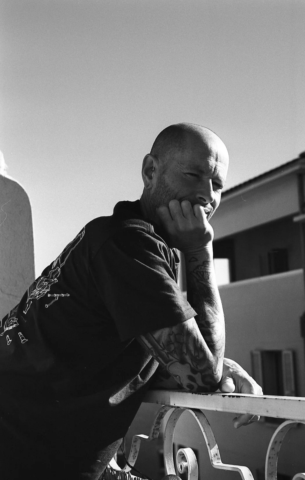 Un homme avec un crâne rasé et des tatouages, pliant le bras, pose avec la main sur son visage, dans un environnement extérieur en noir et blanc.