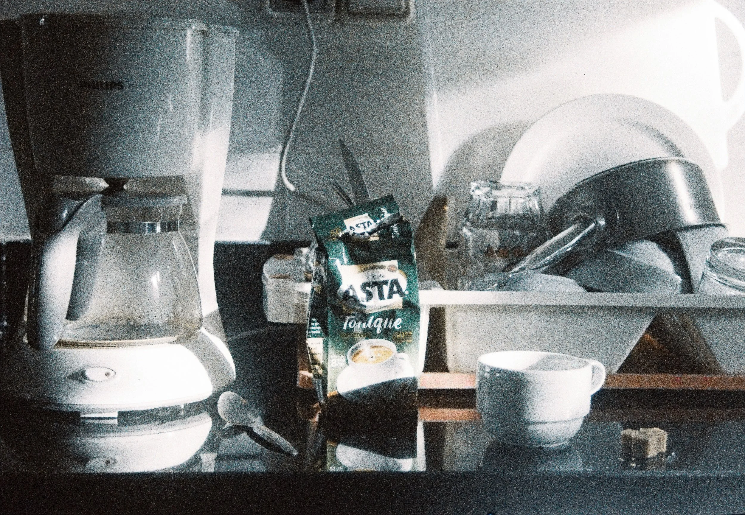 Une cafetière à filtre blanche, un sachet de café instantané, une tasse blanche, un verre et un objet indéterminé sur une surface sombre dans une cuisine