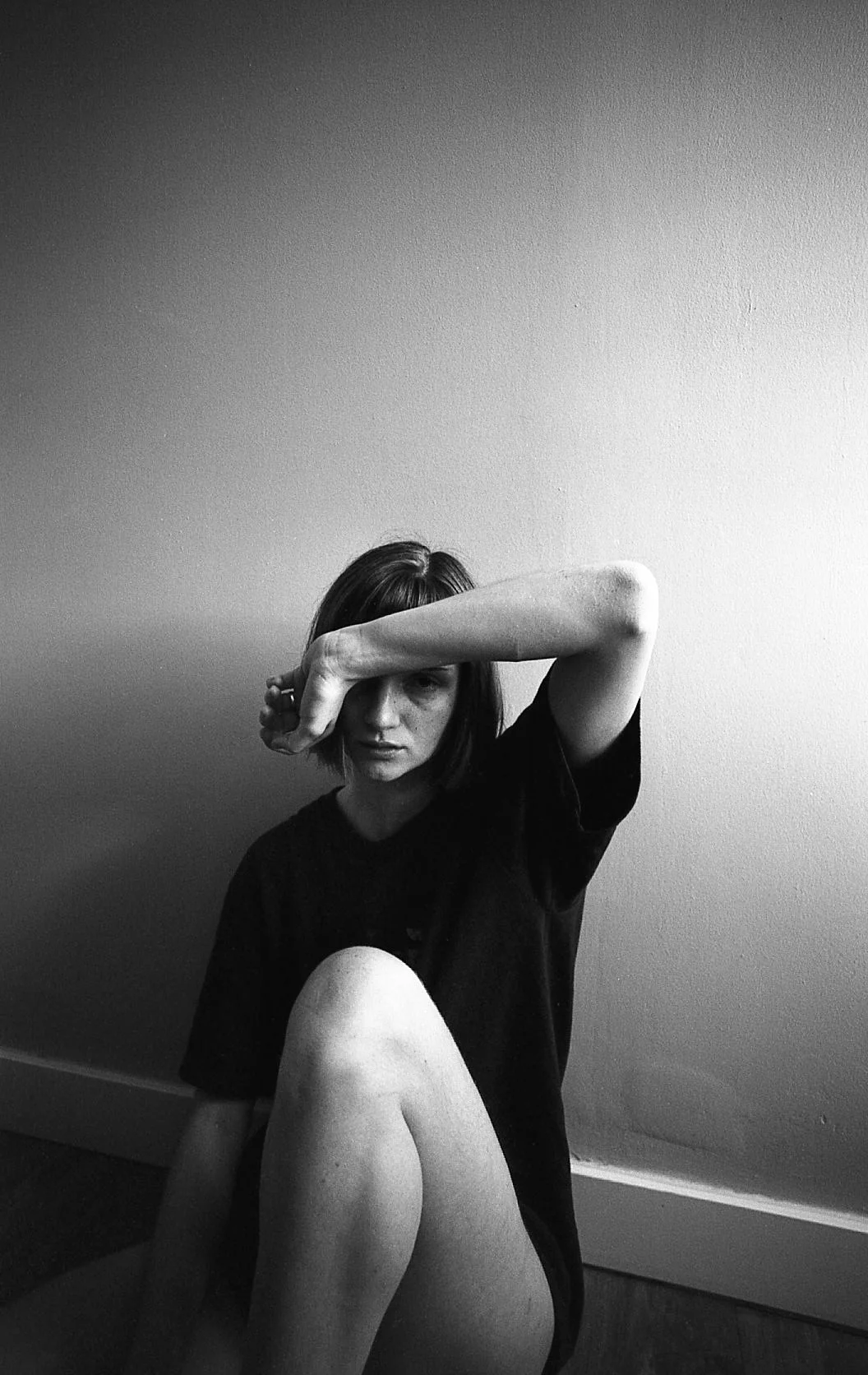 Jeune femme aux cheveux courts, portant un t-shirt noir, assise par terre contre un mur blanc, avec un bras couvrant son visage dans une pose artistique en noir et blanc.