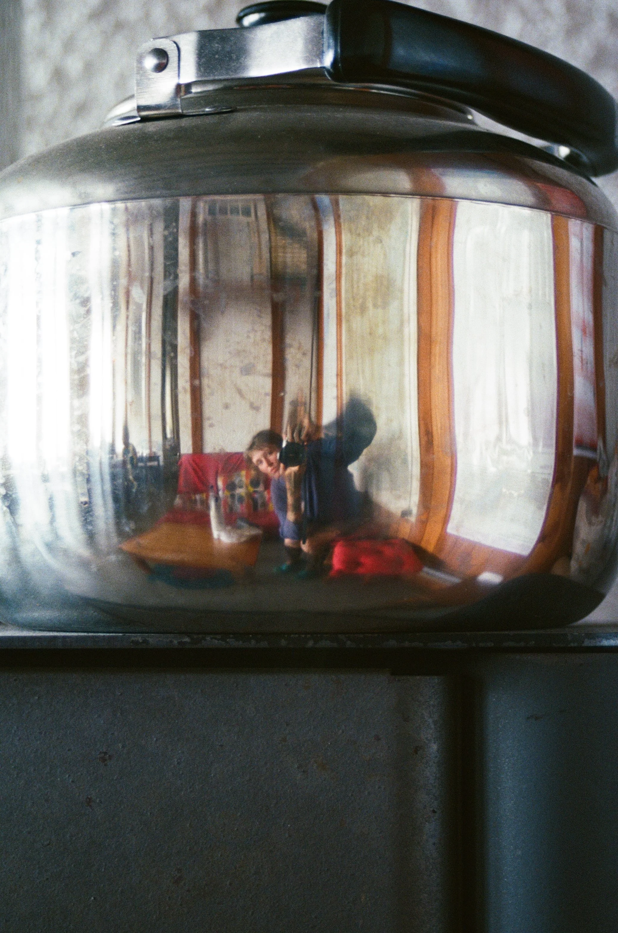 Reflet d'une personne dans une bouilloire en métal posée sur une surface en bois, avec un intérieur en bois et des fenêtres en arrière-plan.