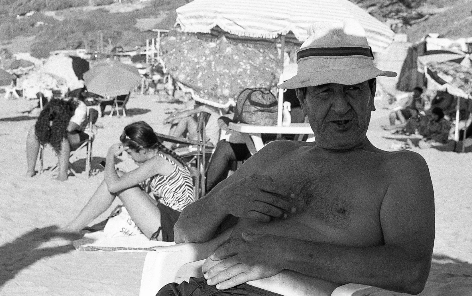 Un homme torse nu portant un chapeau, assis sur une chaise à la plage, avec plusieurs parasols et personnes en arrière-plan.