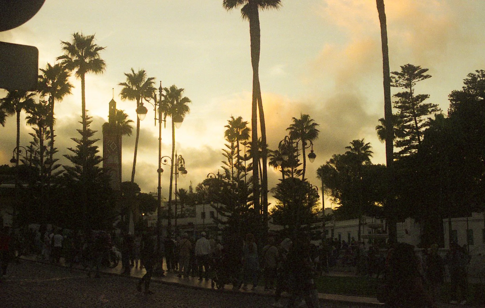 Coucher de soleil avec des palmiers et des lampadaires dans une ville, plusieurs personnes marchent dans la rue.