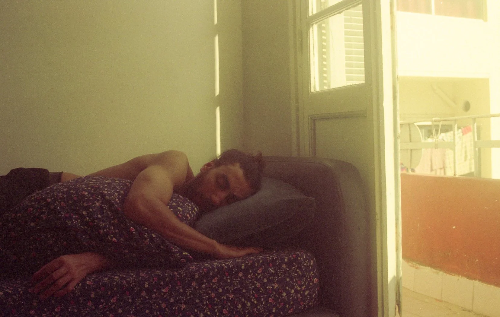 Un homme dort sur un canapé, dans une pièce avec une porte-fenêtre menant à un balcon ensoleillé.