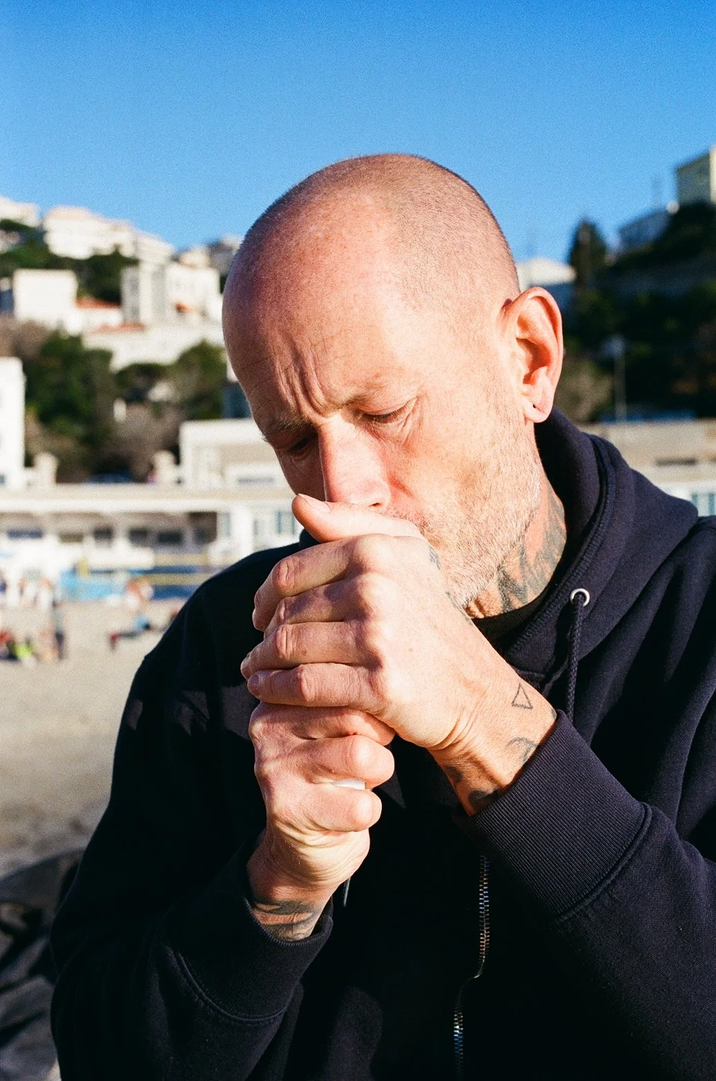 Un homme chauve prie ou médite sur la plage avec des bâtiments en arrière-plan ensoleillé.