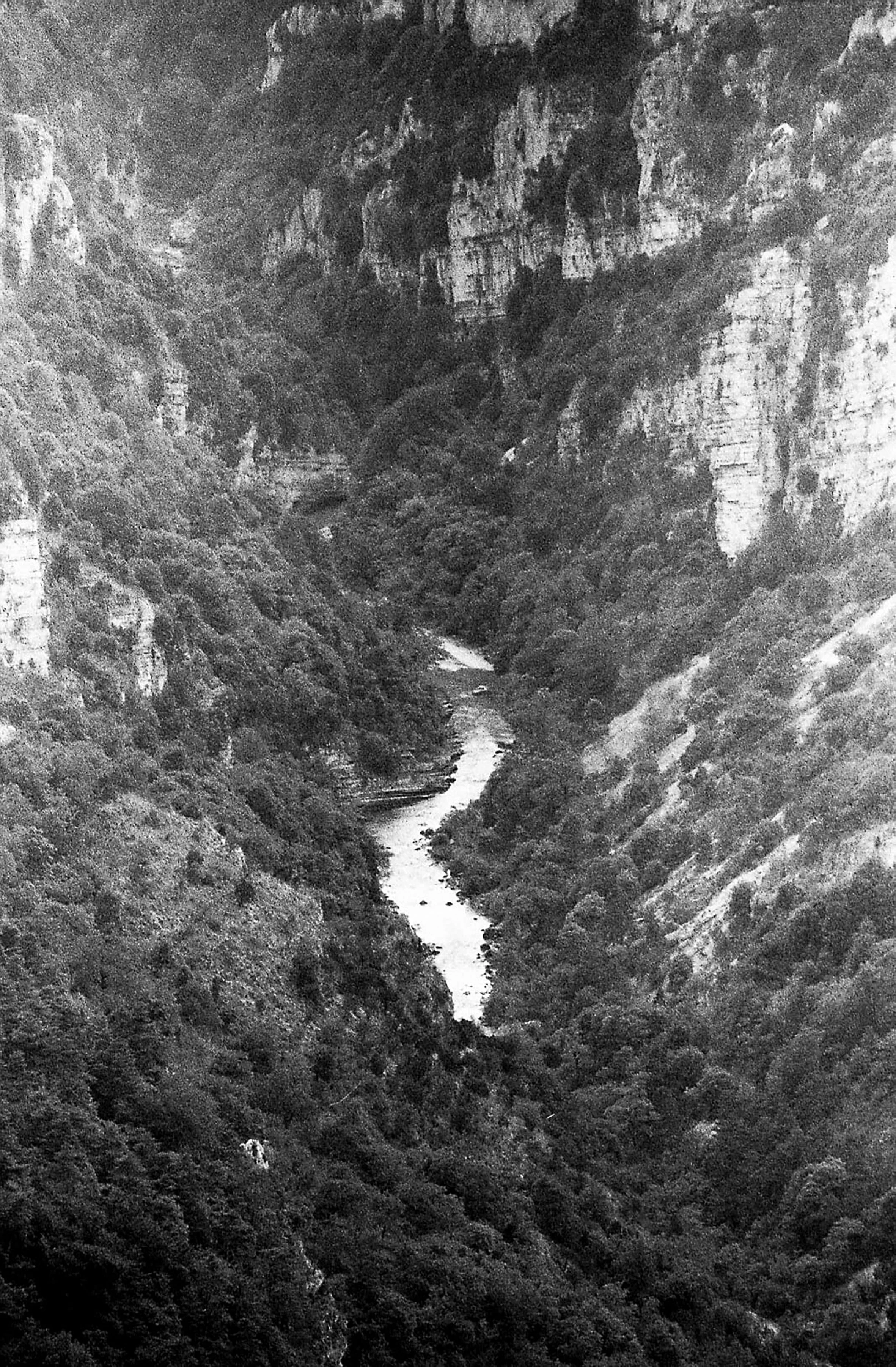 Photo en noir et blanc montrant une vallée profonde avec une rivière serpentant au fond, entourée de hautes falaises rocheuses et de végétation dense.