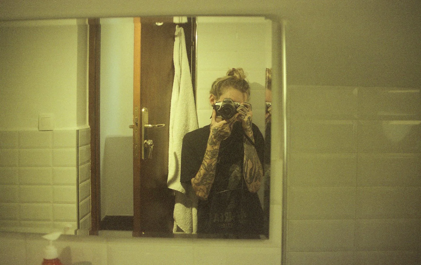 Une personne prenant une photo dans un miroir, avec des tatouages visibles sur les bras, se tenant dans une pièce avec une porte en arrière-plan et un linge blanc suspendu.