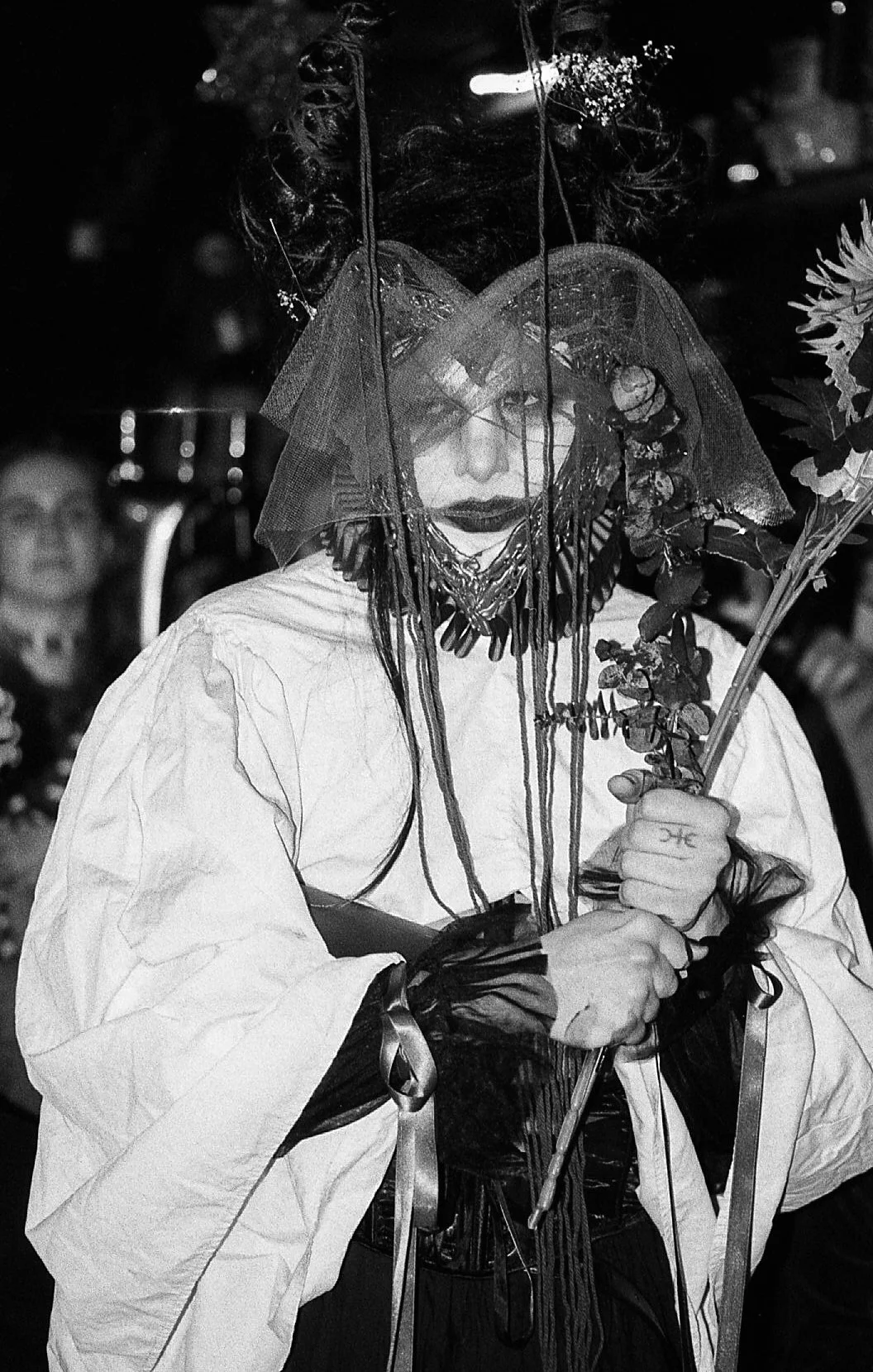 Personne portant un déguisement sombre avec un voile en tulle, des accessoires sombres et un bouquet de fleurs, dans un style gothique ou artistique, lors d'un événement ou d'une cérémonie.