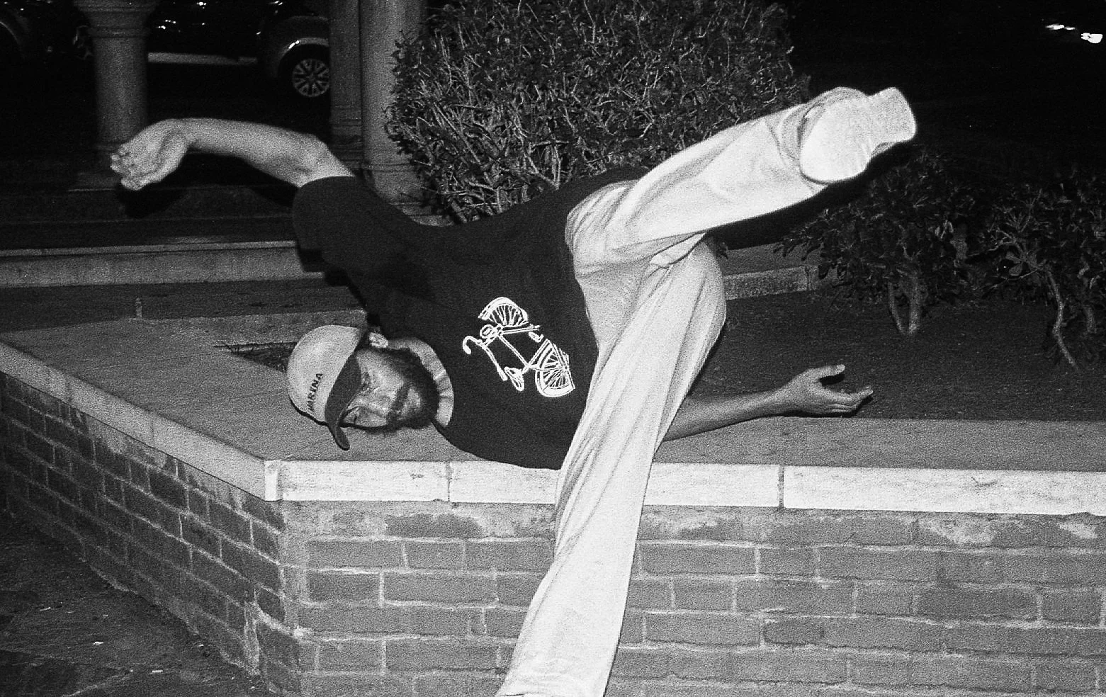 Un homme portant un sweat noir avec un logo de vélo, un pantalon à rayures, une casquette blanche, et des lunettes, est assis par terre contre un muret en brique, dans une zone extérieure, de nuit.