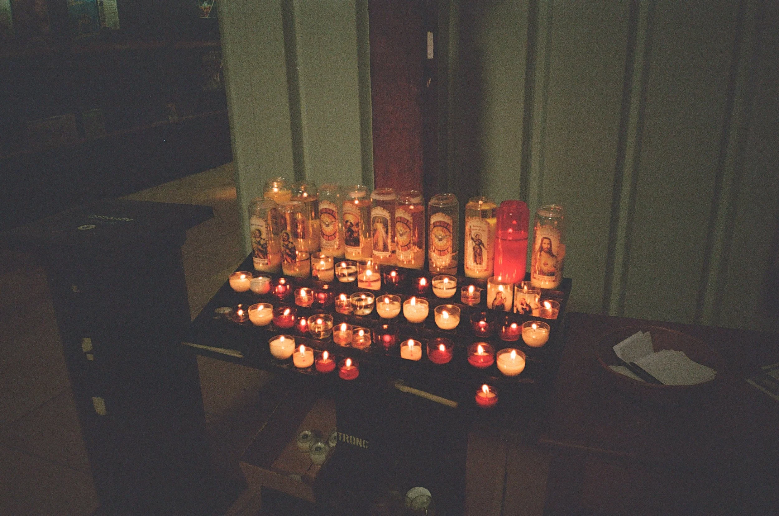 Un panneau avec plusieurs bougies allumées, certaines en verre, et des images religieuses. La scène est sombre, évoquant une atmosphère de recueillement ou de prière.