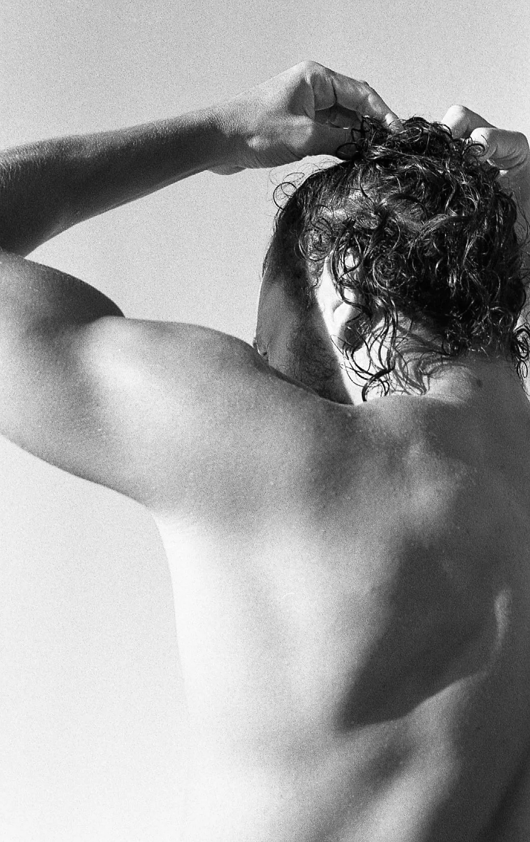 Un homme nu vu de dos, avec une main dans ses cheveux, en noir et blanc.