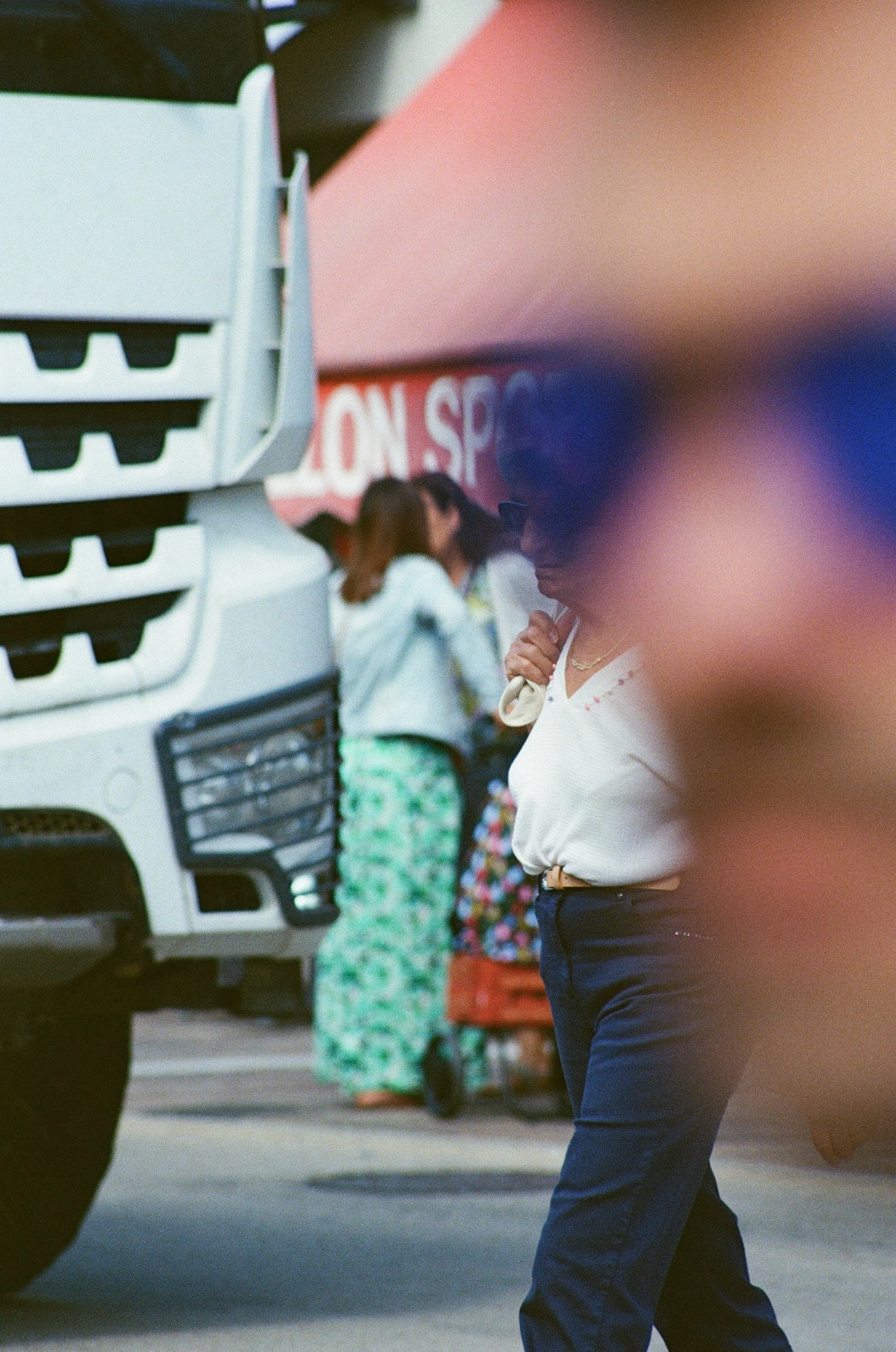 Une femme portant des lunettes de soleil, un chemisier blanc et un pantalon foncé, se tient près d'un camion blanc dans une rue animée, avec d'autres personnes en arrière-plan.