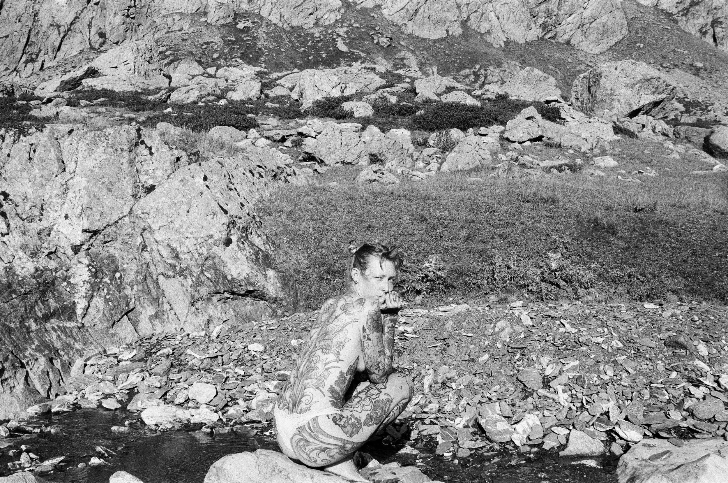 Femme aux cheveux blonds et tatouages, assise sur des rochers près d'une rivière ou d'un ruisseau dans un paysage de montagne rocheuse.