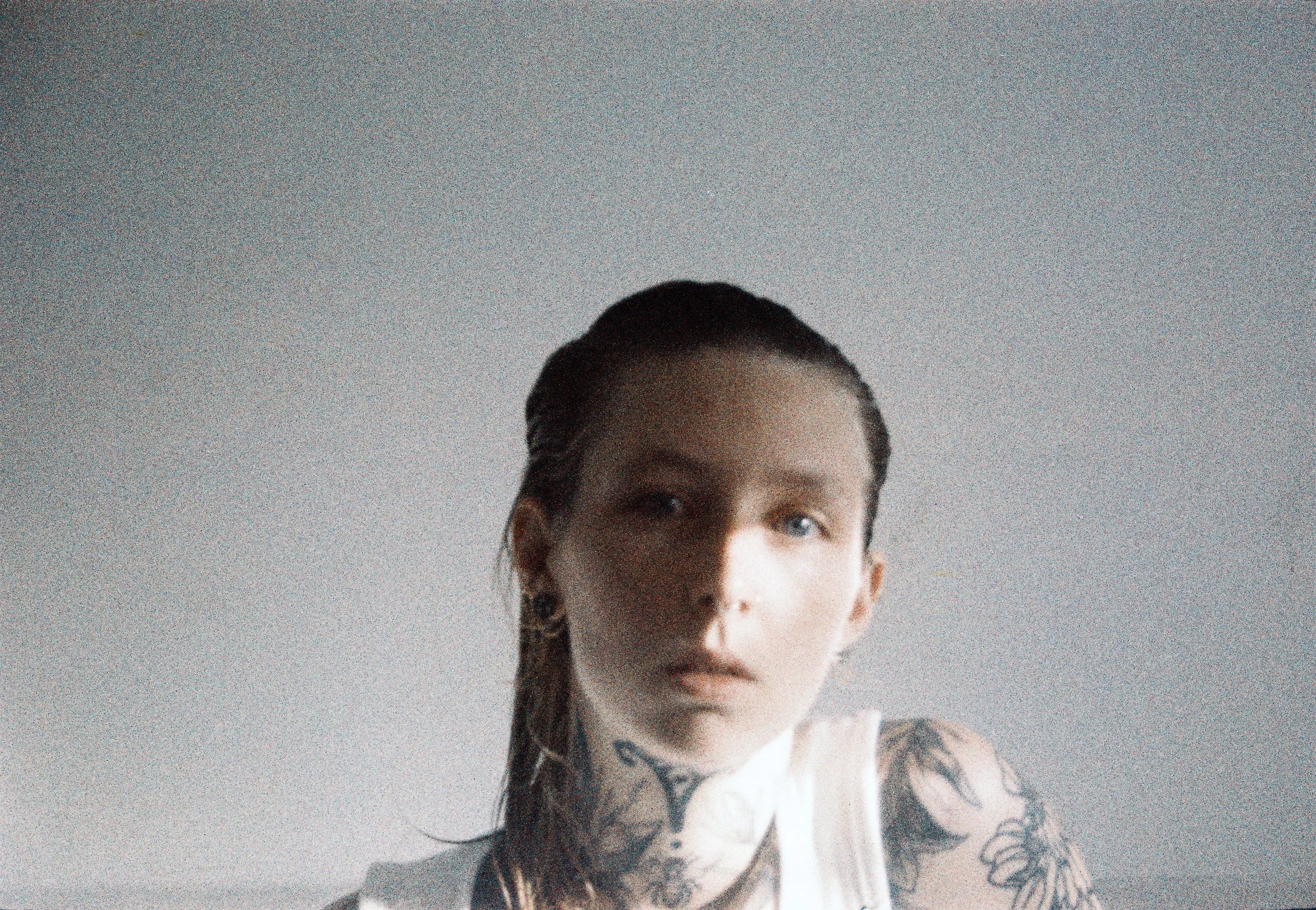 Portrait d'une femme avec tatouages sur le bras et le cou, regardant vers l'objectif, fond gris clair.