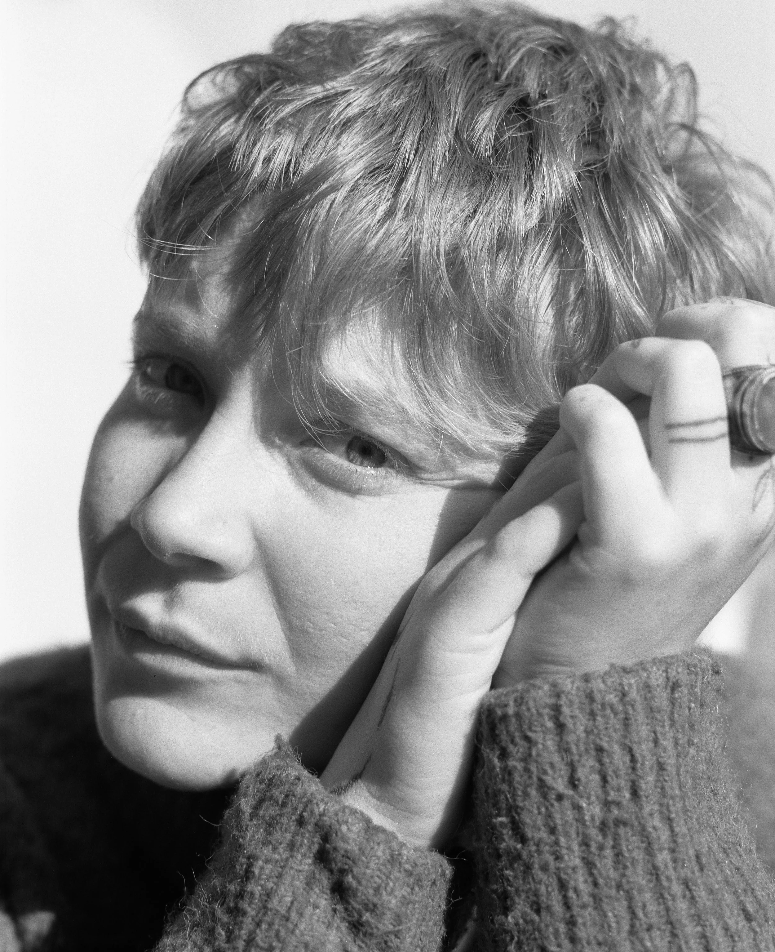 Un portrait en noir et blanc d'une personne avec des cheveux courts et bouclés, tenant sa main près du visage, portant un vêtement en laine.