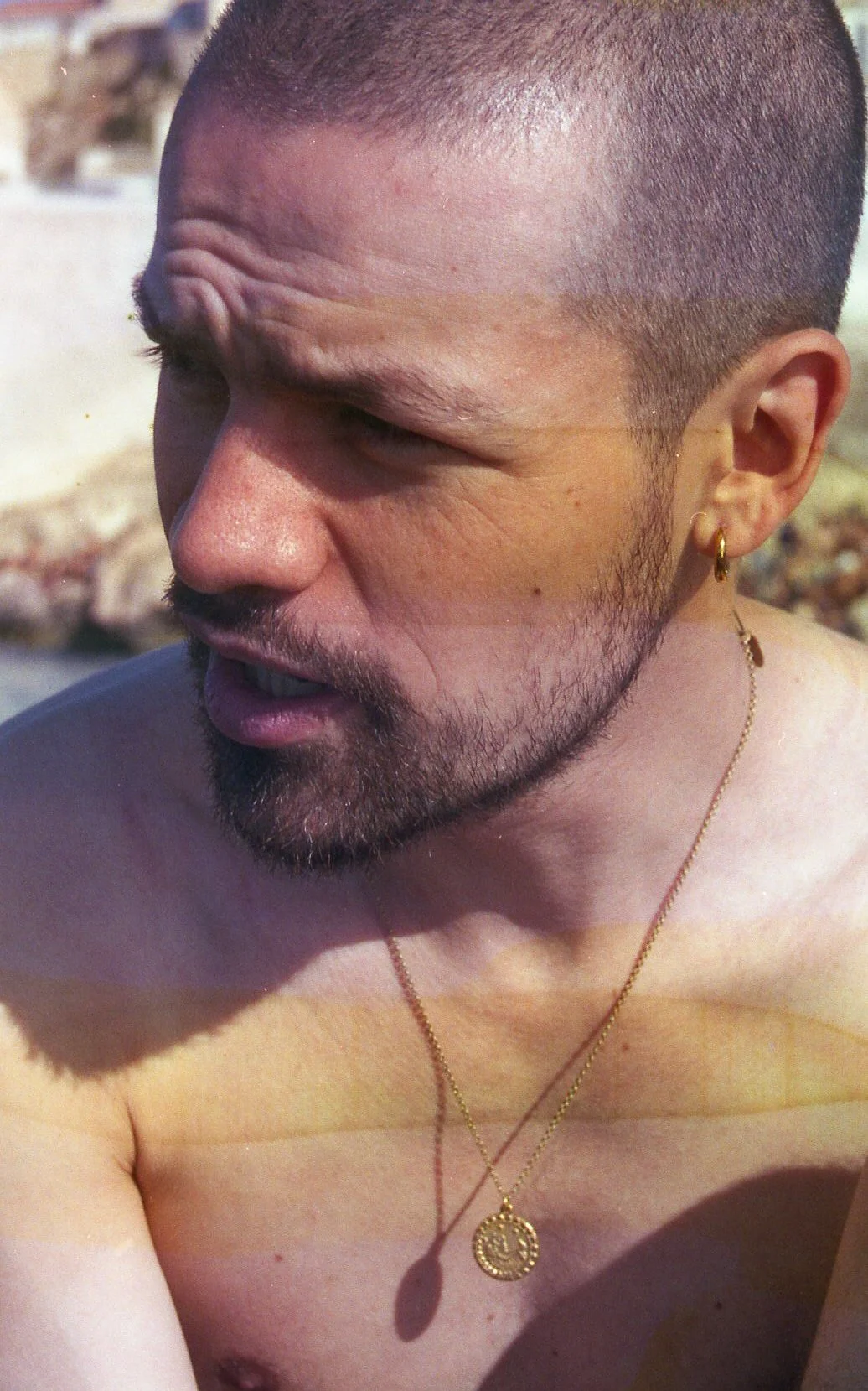 Un homme chauve avec une barbe, portant une boucle d'oreille en or, un collier en chaîne avec un pendentif rond, en extérieur avec un fond rocheux.