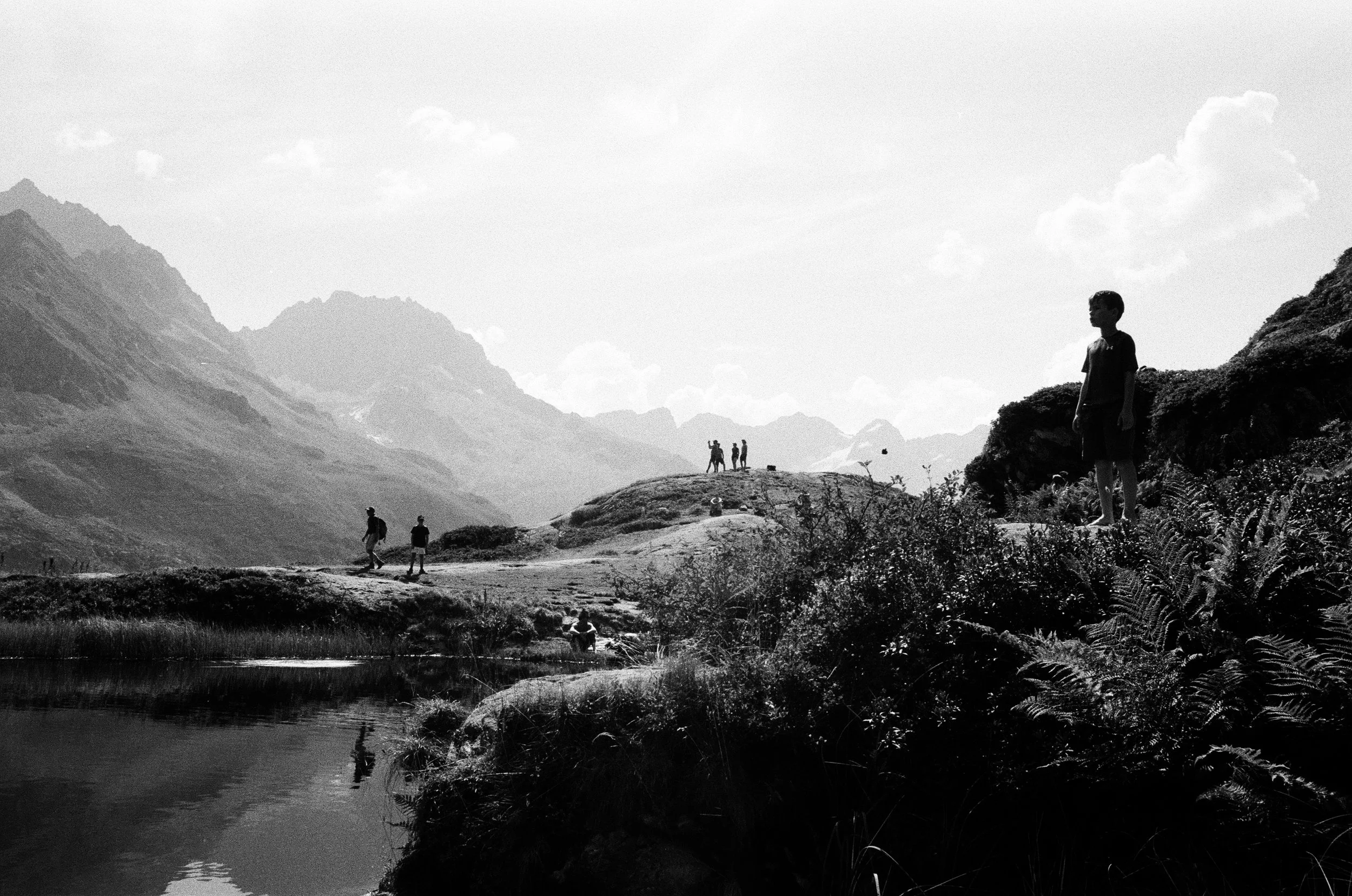 Paysage montagneux avec des randonneurs, un homme en premier plan sur un rocher, et un groupe de personnes au loin, près d'un lac, en images en noir et blanc.