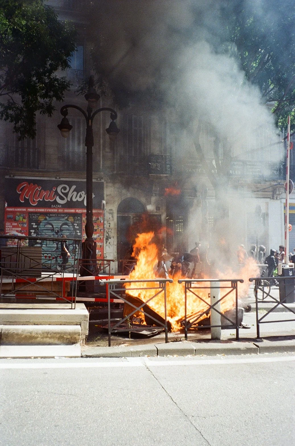 Un feu dans une poubelle en plein air, avec de la fumée qui s'élève, en face d'un bâtiment de commerce avec une enseigne 'Mini Shop', et des personnes qui semblent réagir à l'incident.