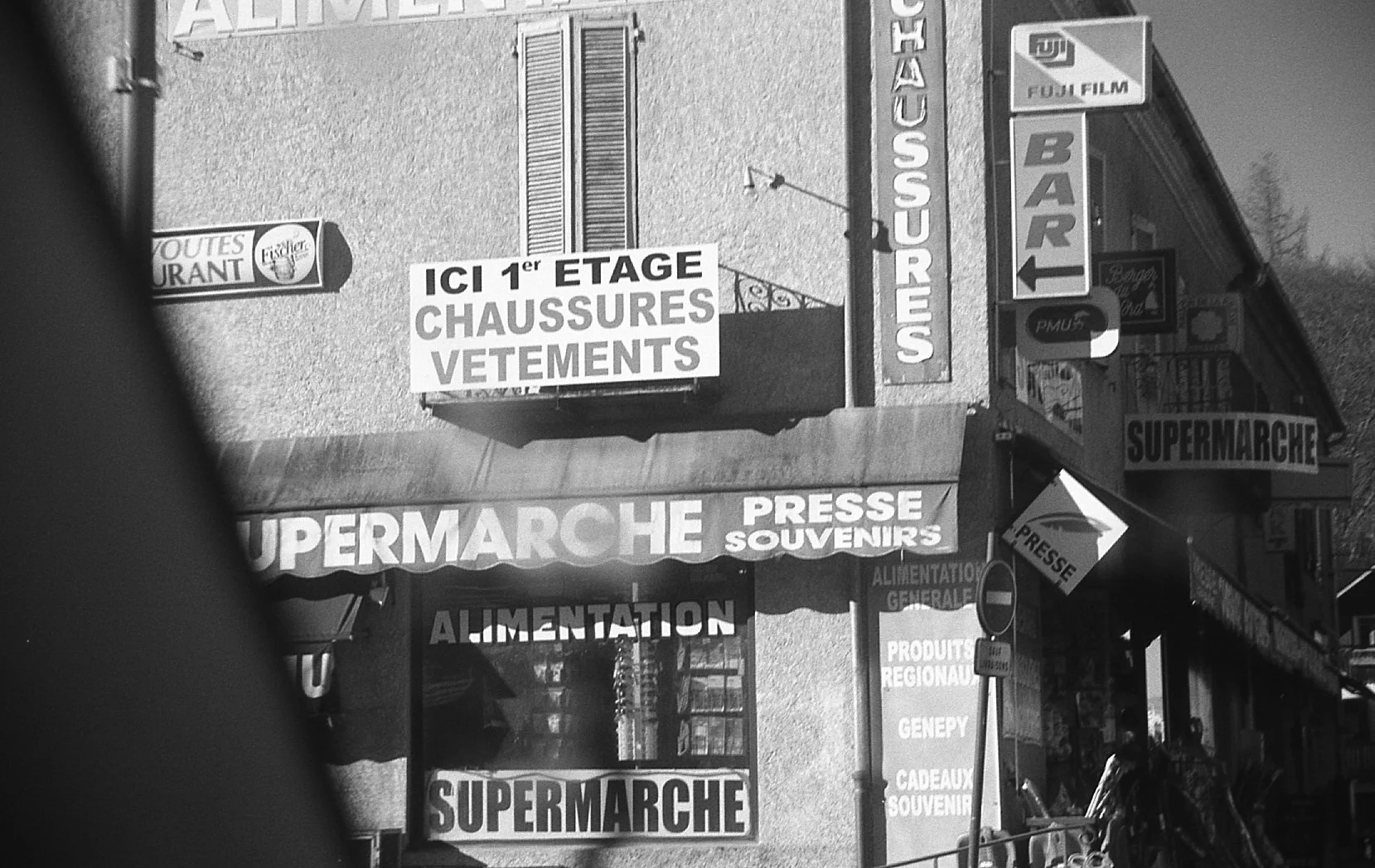 Une rue avec plusieurs enseignes en français pour un supermarché, un magasin de chaussures, vêtements, et souvenirs, avec des panneaux indiquant le premier étage et des produits alimentaires.