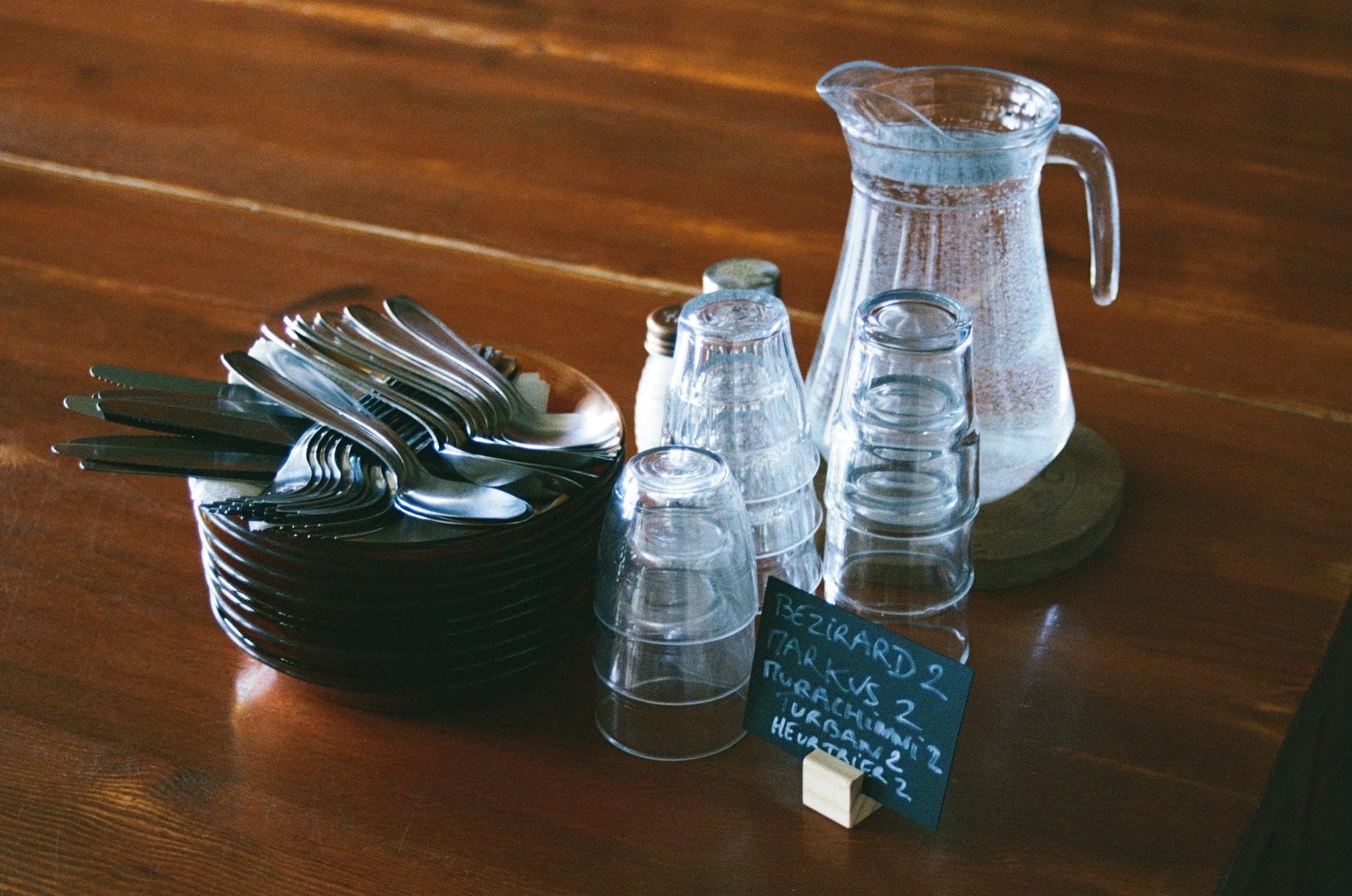 Verres en plastique empilés, carafe d'eau, assiette avec des couverts en métal, petites piques, et une petite ardoise avec une liste de noms.