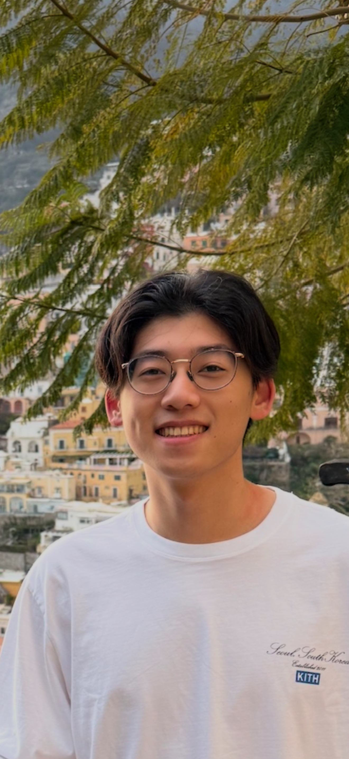 A young man with glasses smiling outdoors with a pink and yellow hillside cityscape in the background and green overhanging tree branches above.