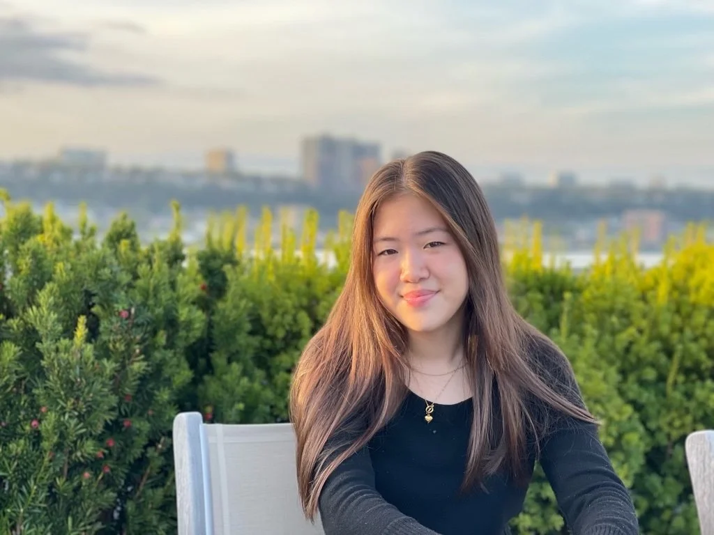 A young woman with long brown hair smiling while sitting outdoors at a table, with green bushes and a cityscape in the background during daylight.
