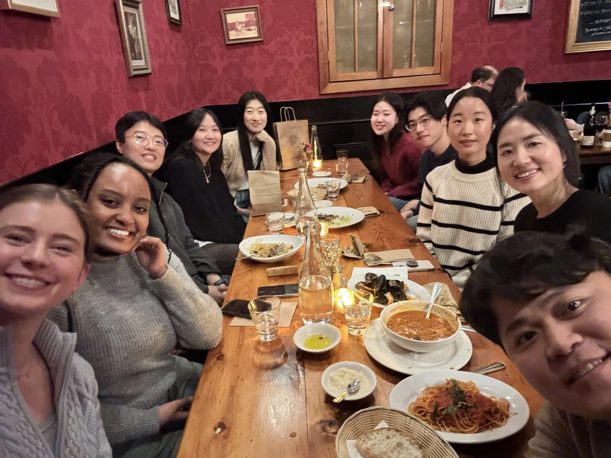 A group of ten people sitting around a long wooden table at a restaurant, smiling at the camera, with plates of food including pasta and soup, and drinks on the table.