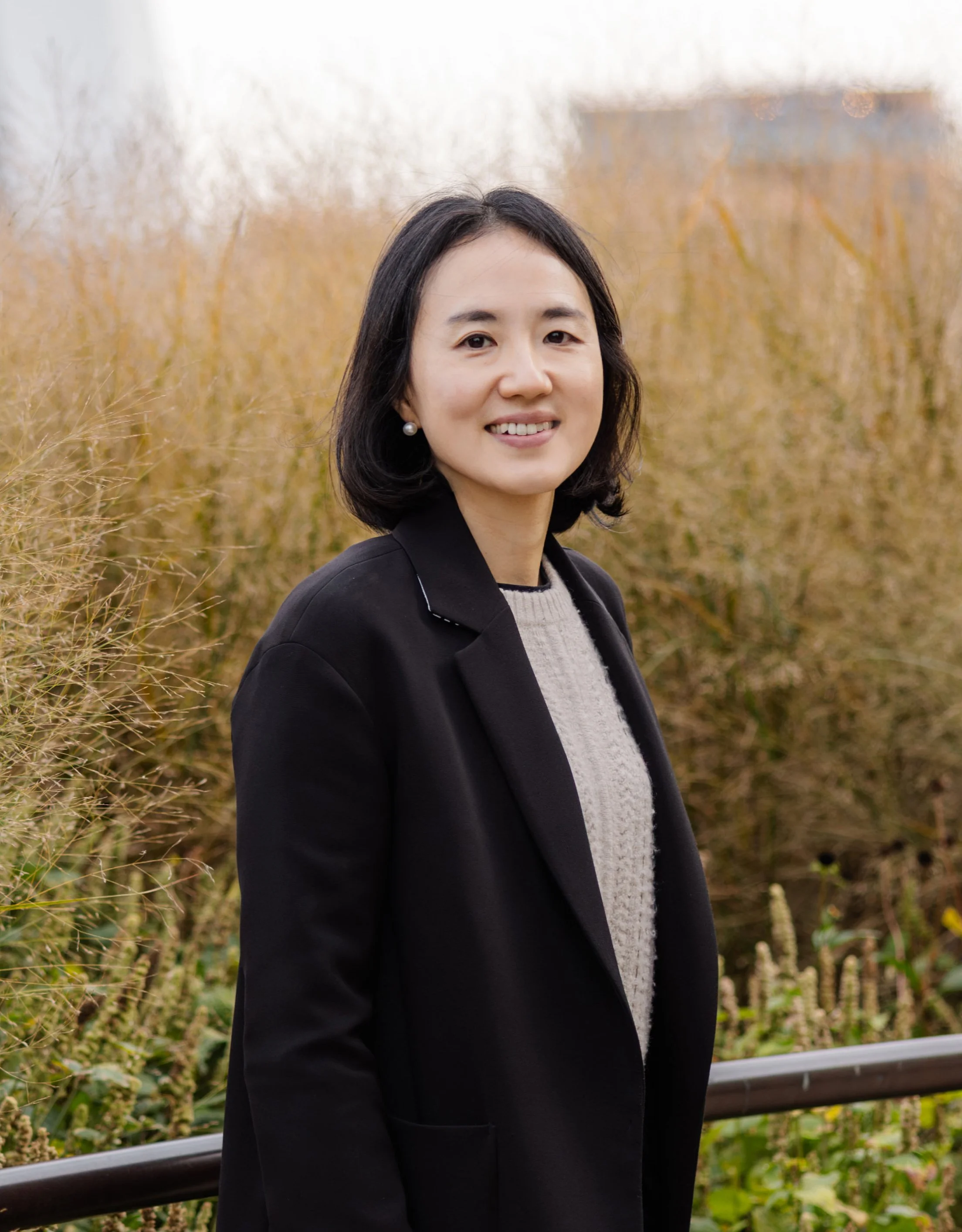 A woman with shoulder-length black hair, dressed in a black blazer and light-colored sweater, stands outdoors in front of tall, dry grasses, smiling at the camera.
