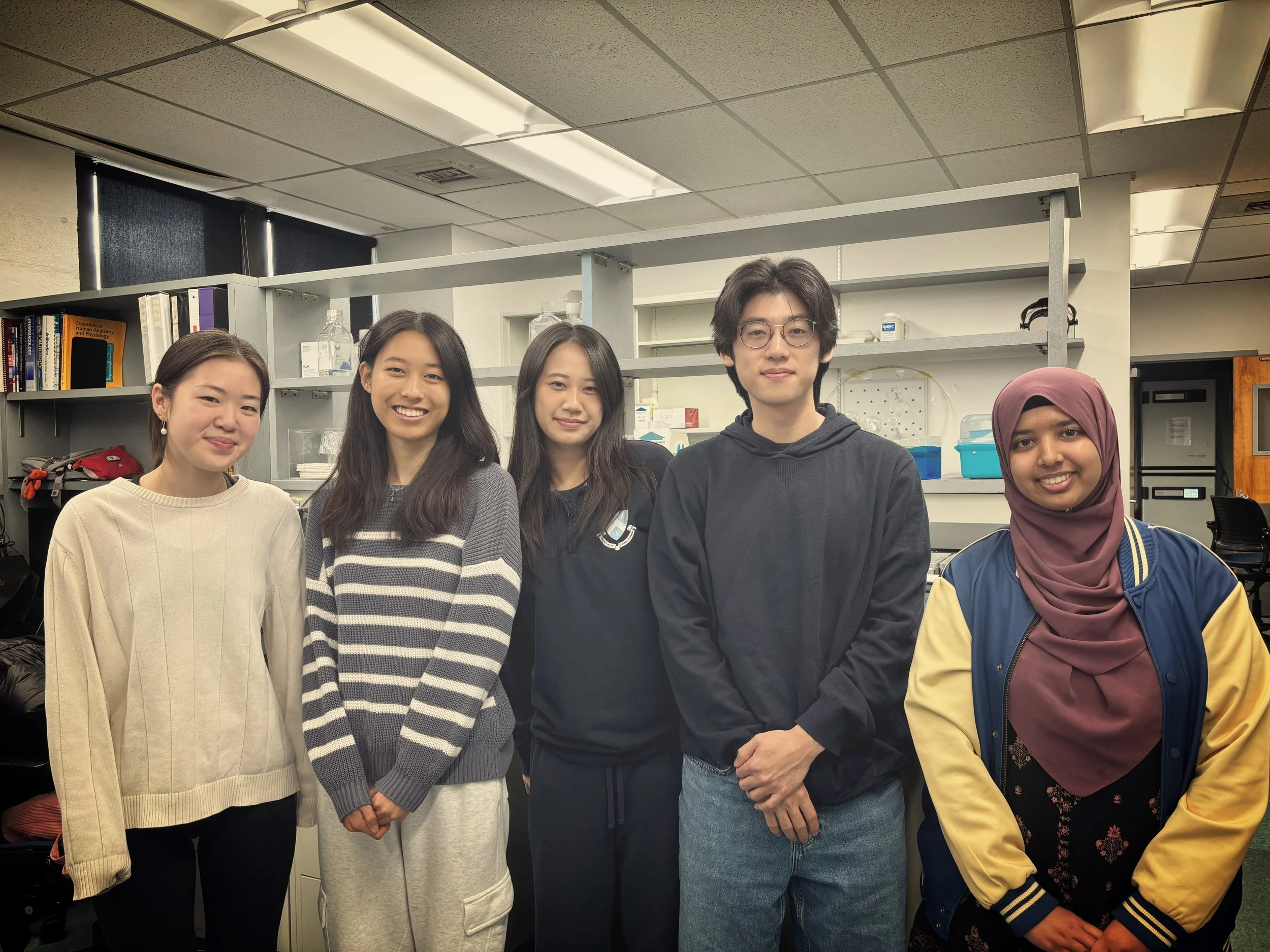 Group of five young adults standing in a laboratory or classroom, smiling at the camera, with shelves, books, and laboratory equipment behind them.