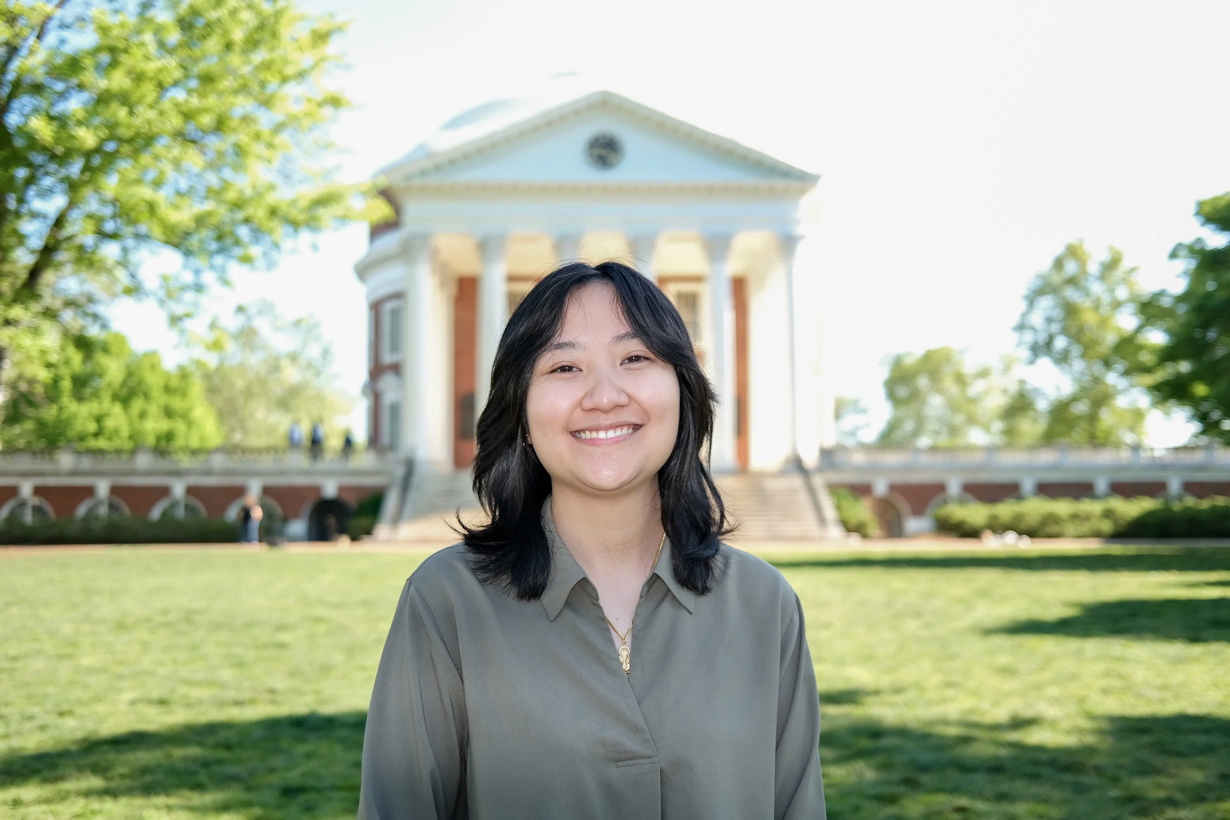 Young woman smiling outdoors on a sunny day with a historic white and red-brick building with columns in the background.
