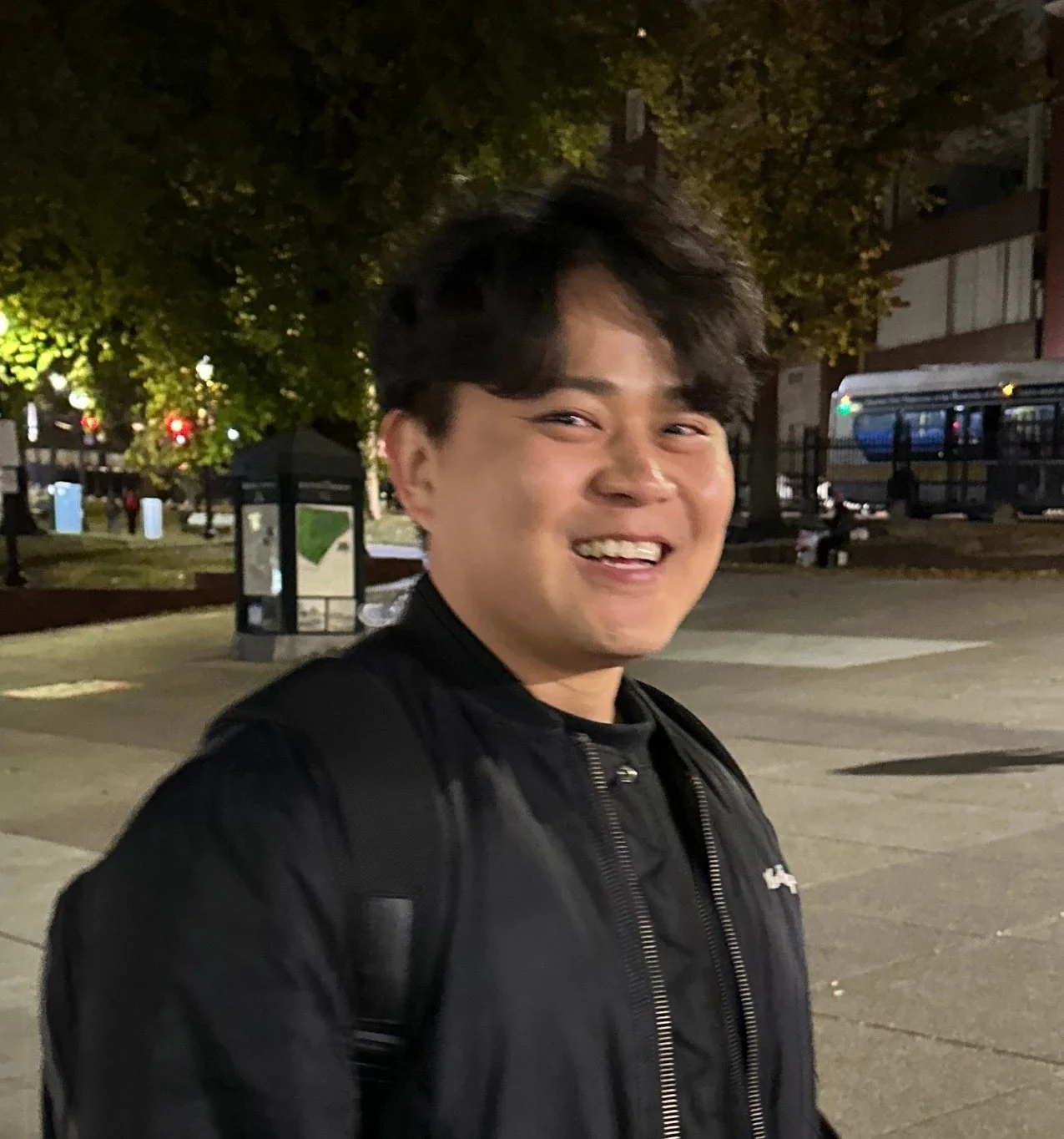 A young man smiling outdoors at night with trees, benches, and a bus in the background.