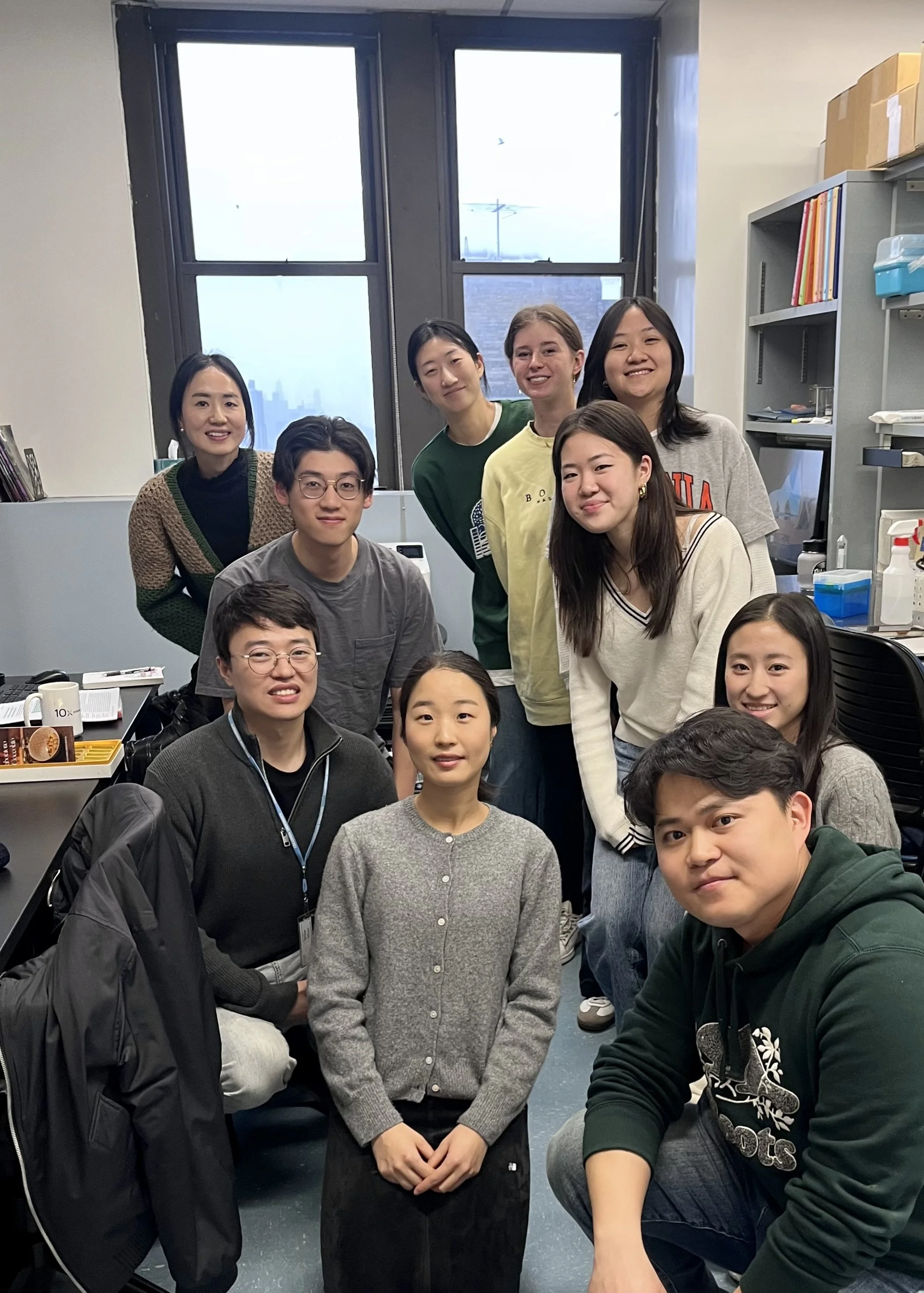 Group of eleven young Asian and Caucasian adults in an office, smiling and posing for a photo with desks, shelves, and windows in the background.