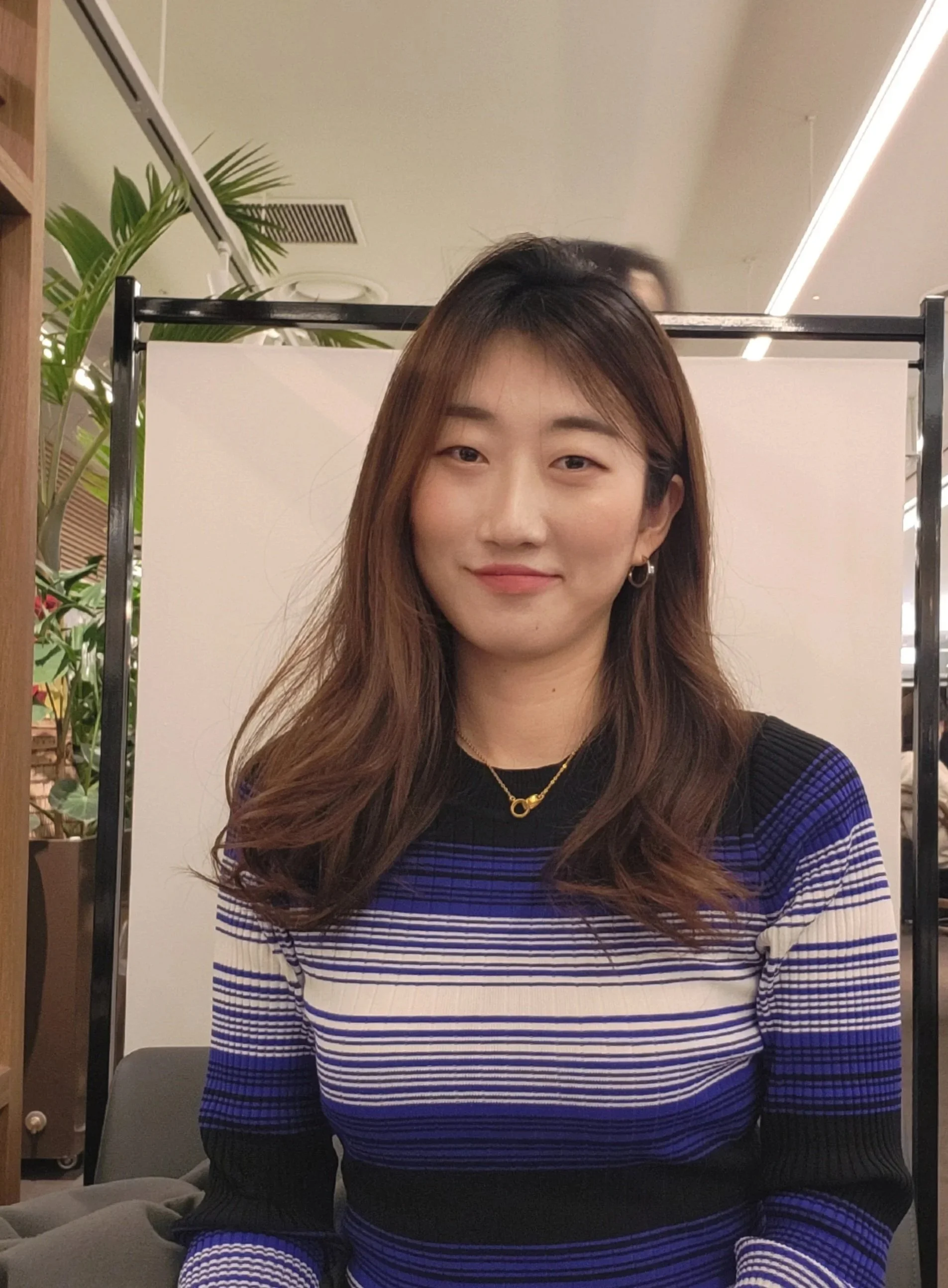 A young woman with long brown hair and a slight smile, wearing a striped blue, white, and black sweater, a gold necklace, and hoop earrings, seated indoors with a white partition and green plants in the background.